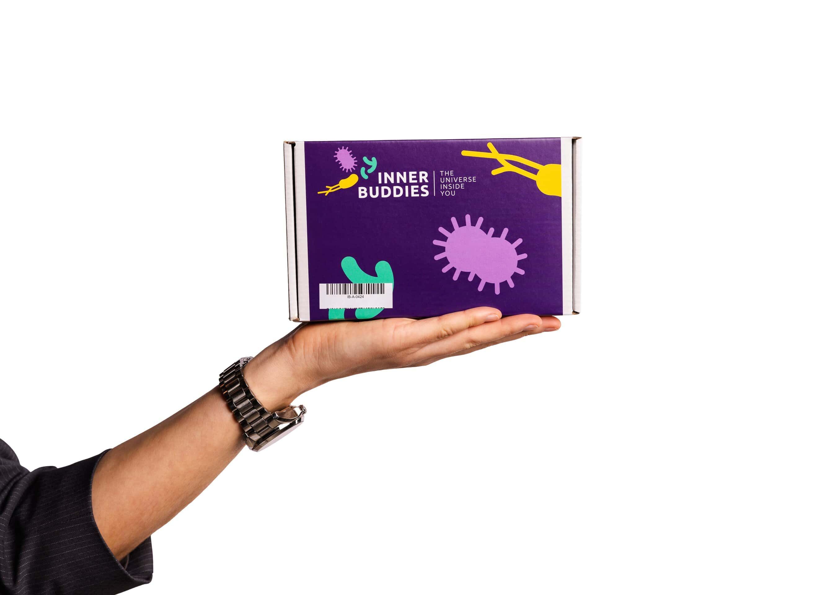 Inner Buddies Mikrobiom Testkit-Verpackung, gehalten in einer Hand, mit bunten Mikroben-Illustrationen.