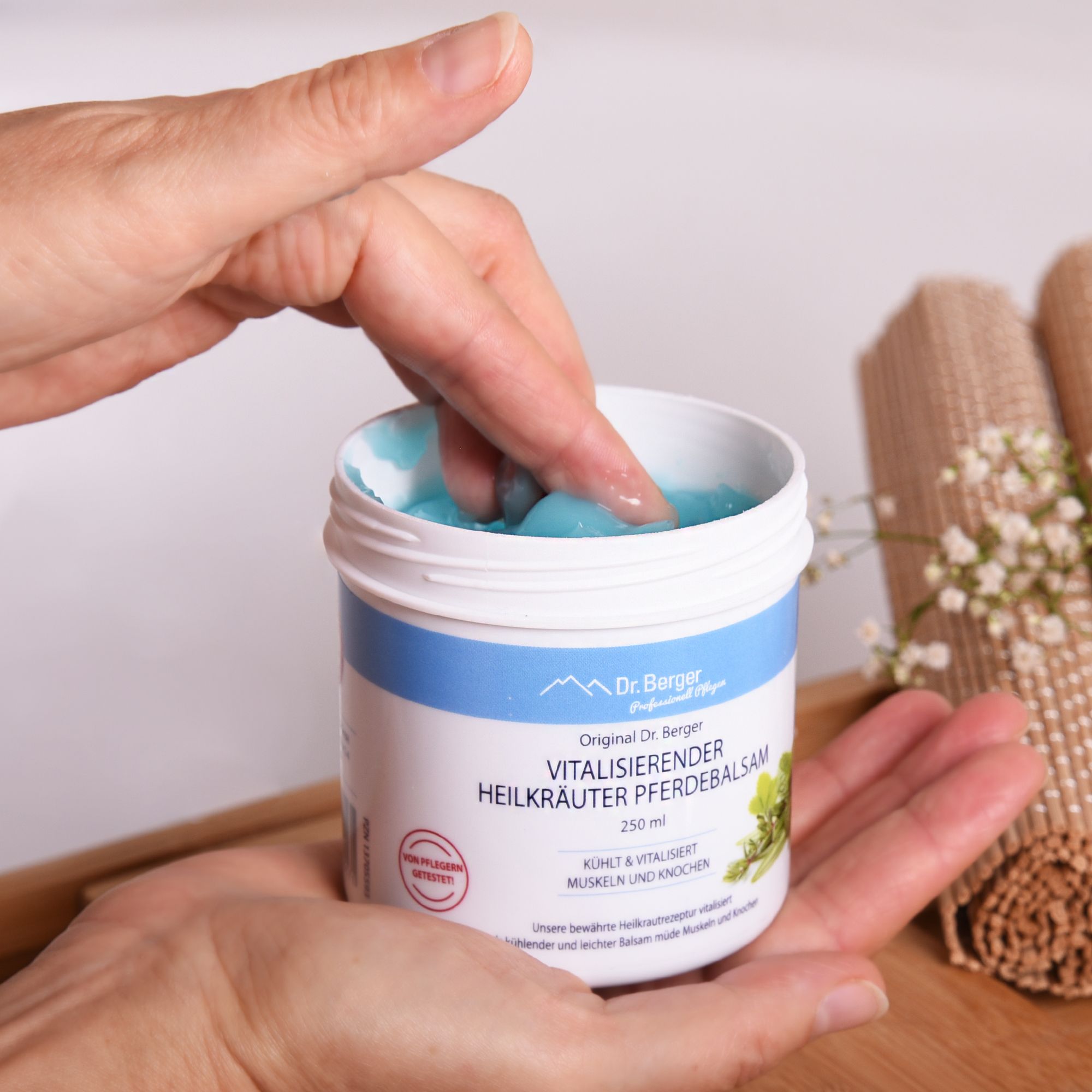 Hände entnehmen blauen Balsam aus geöffnetem weißen Tiegel. Produktbezeichnung sichtbar. Hintergrund: Handtücher.