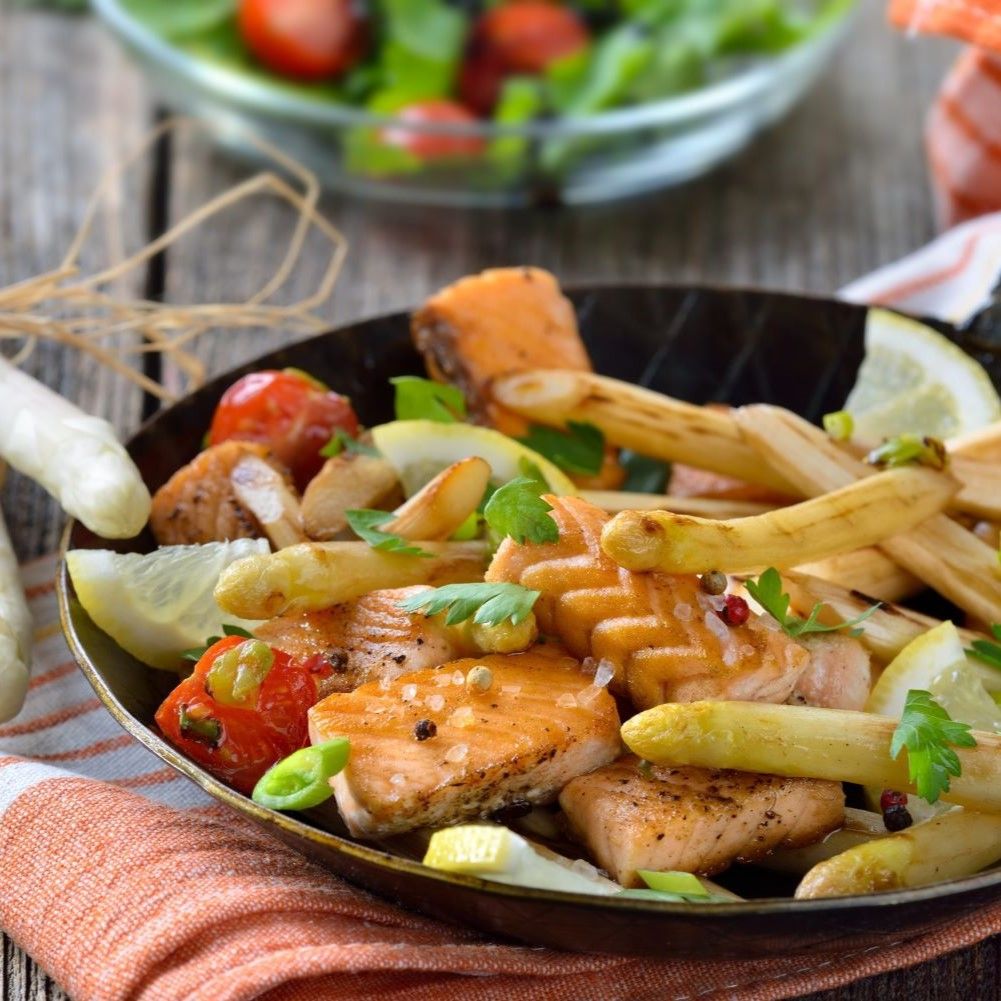 Gebratener Lachs mit Spargel, Tomaten und Zitrone in einer Pfanne. Auf einem Holztisch.