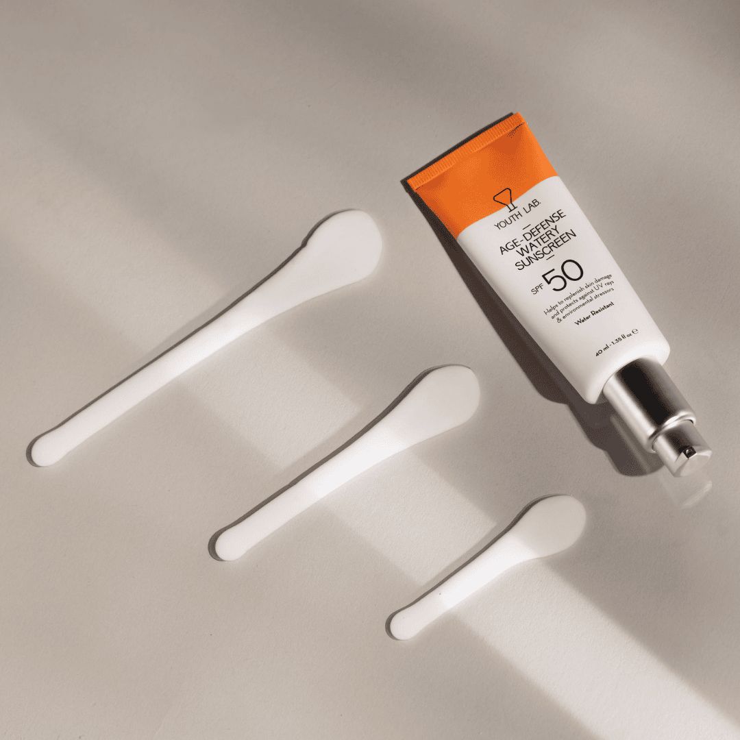 YOUTH LAB Age Defense Watery Sunscreen SPF50 Tube mit Spateln. Weißer Hintergrund, Produkt und Zubehör.