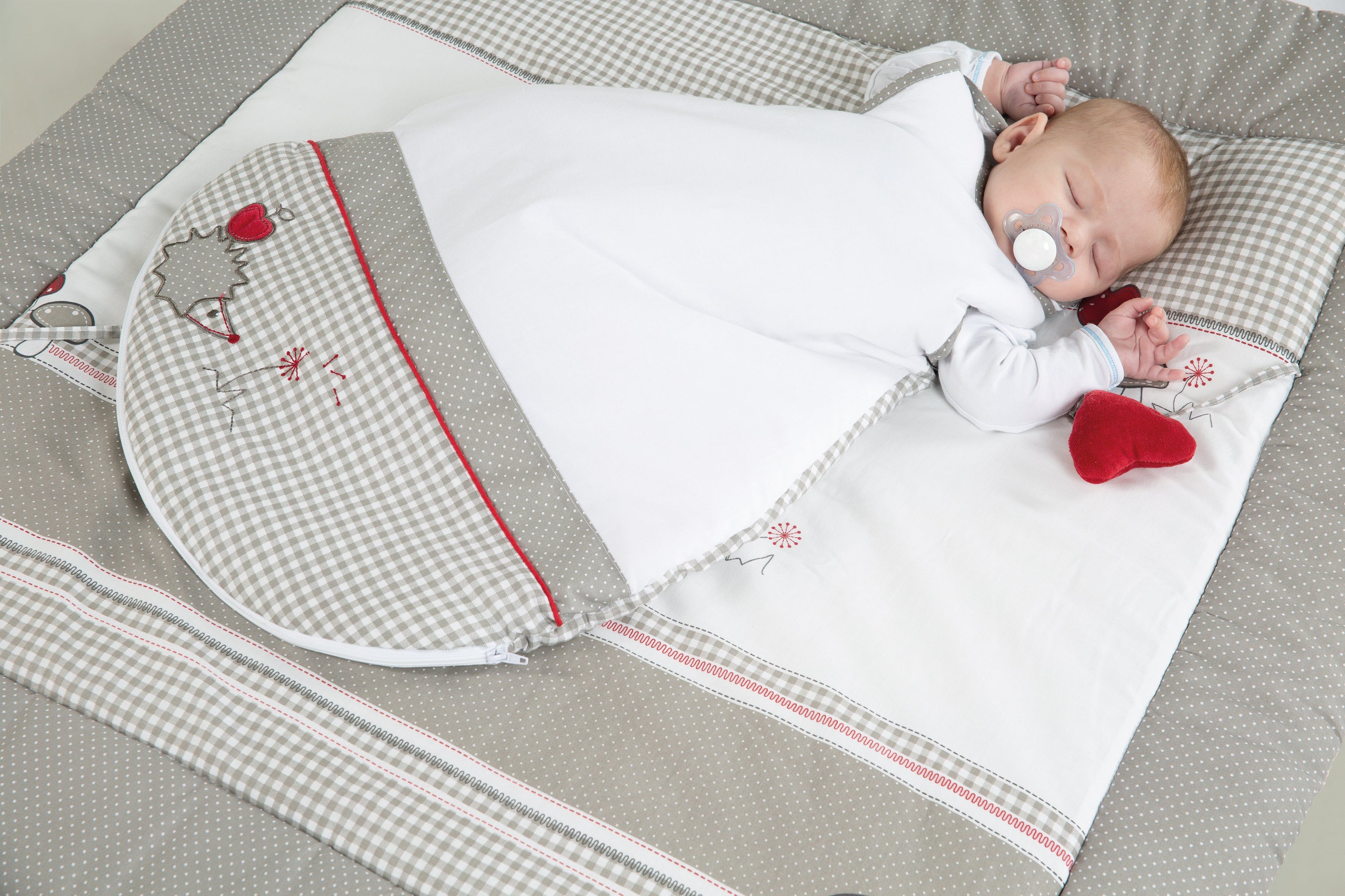 Baby schläft in weißem Schlafsack auf Decke. Decke mit Karomuster und roten Details. Schnuller.