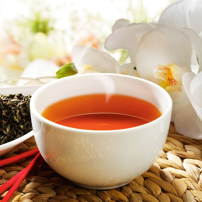 Tasse Tee mit Dampf, neben losem Tee und Orchideen. Der Tee ist bernsteinfarben. Auf einem geflochtenen Untersetzer.