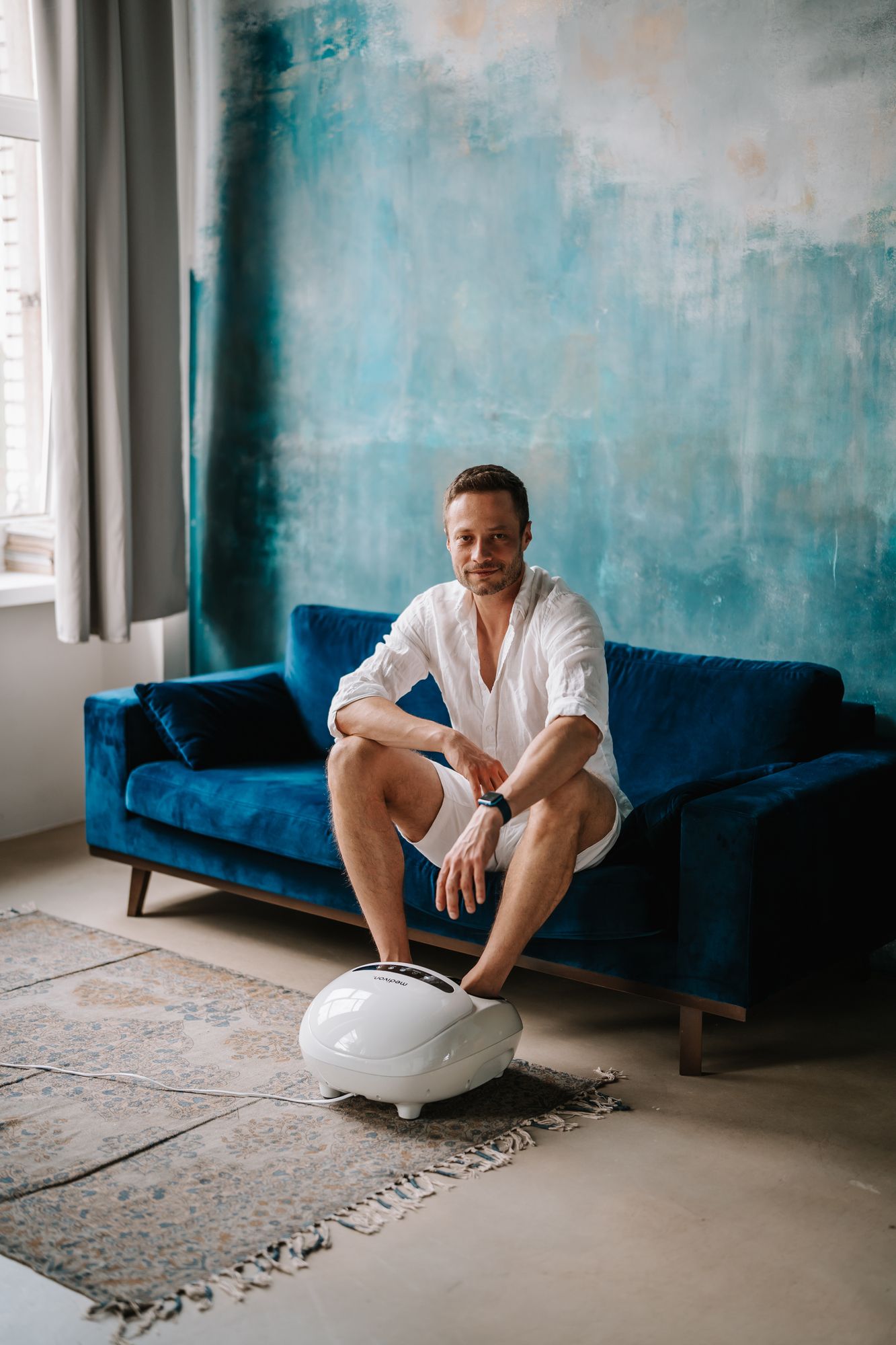 Mann sitzt auf Sofa, Füße in weißem Fußmassagegerät. Blau-grüne Wand im Hintergrund.