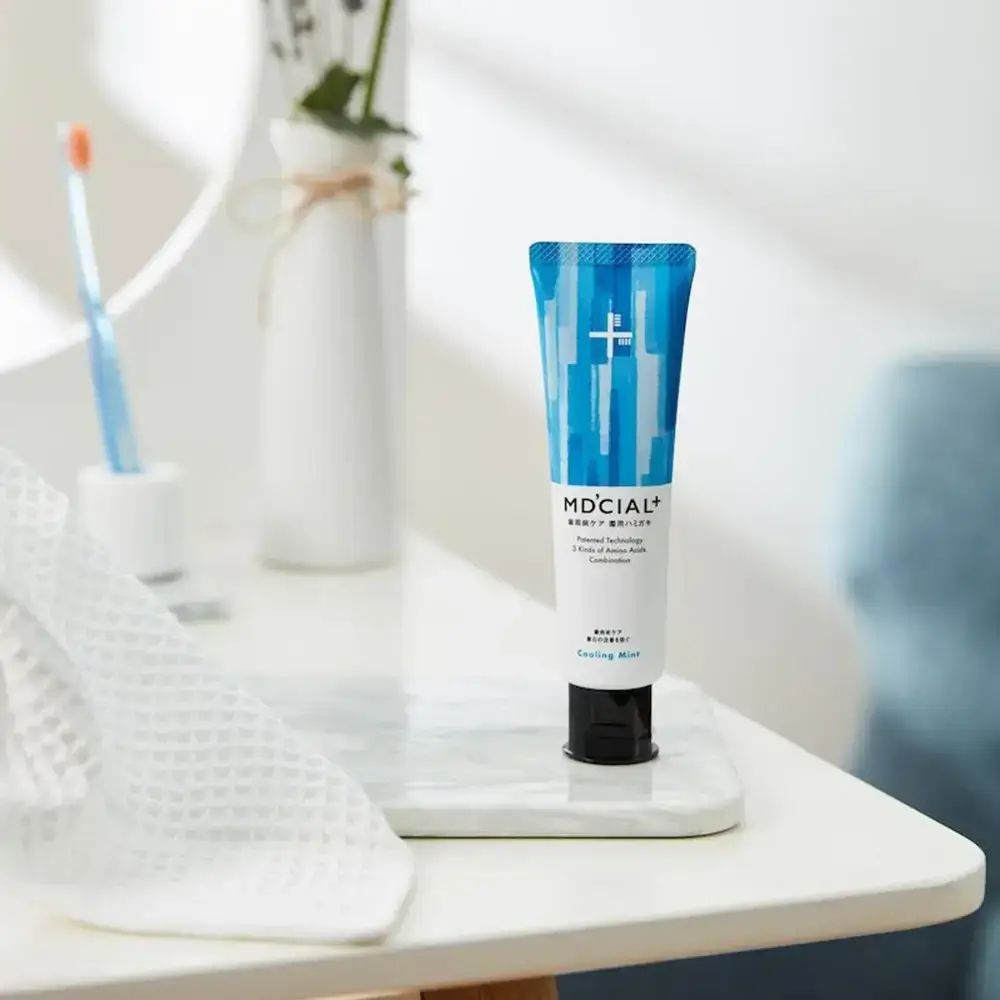 Zahncreme-Tube auf weißer Ablage. Blaue Tube mit weißem Schriftzug. Daneben Zahnbürste und Handtuch.