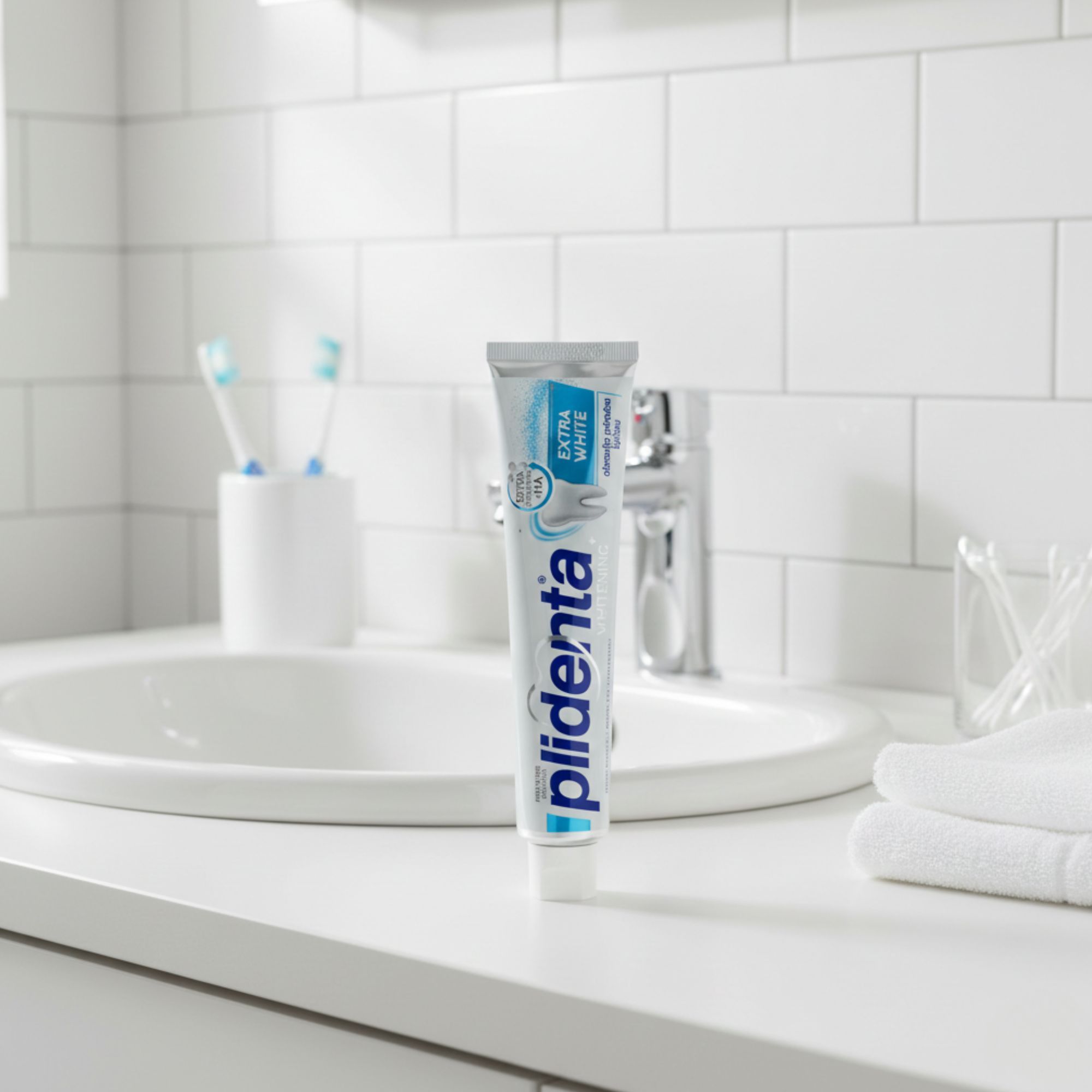 Zahnpasta-Tube auf einem Waschbecken. Aufschrift: Plidenta Extra White. Im Hintergrund: Zahnbürsten und Handtücher.