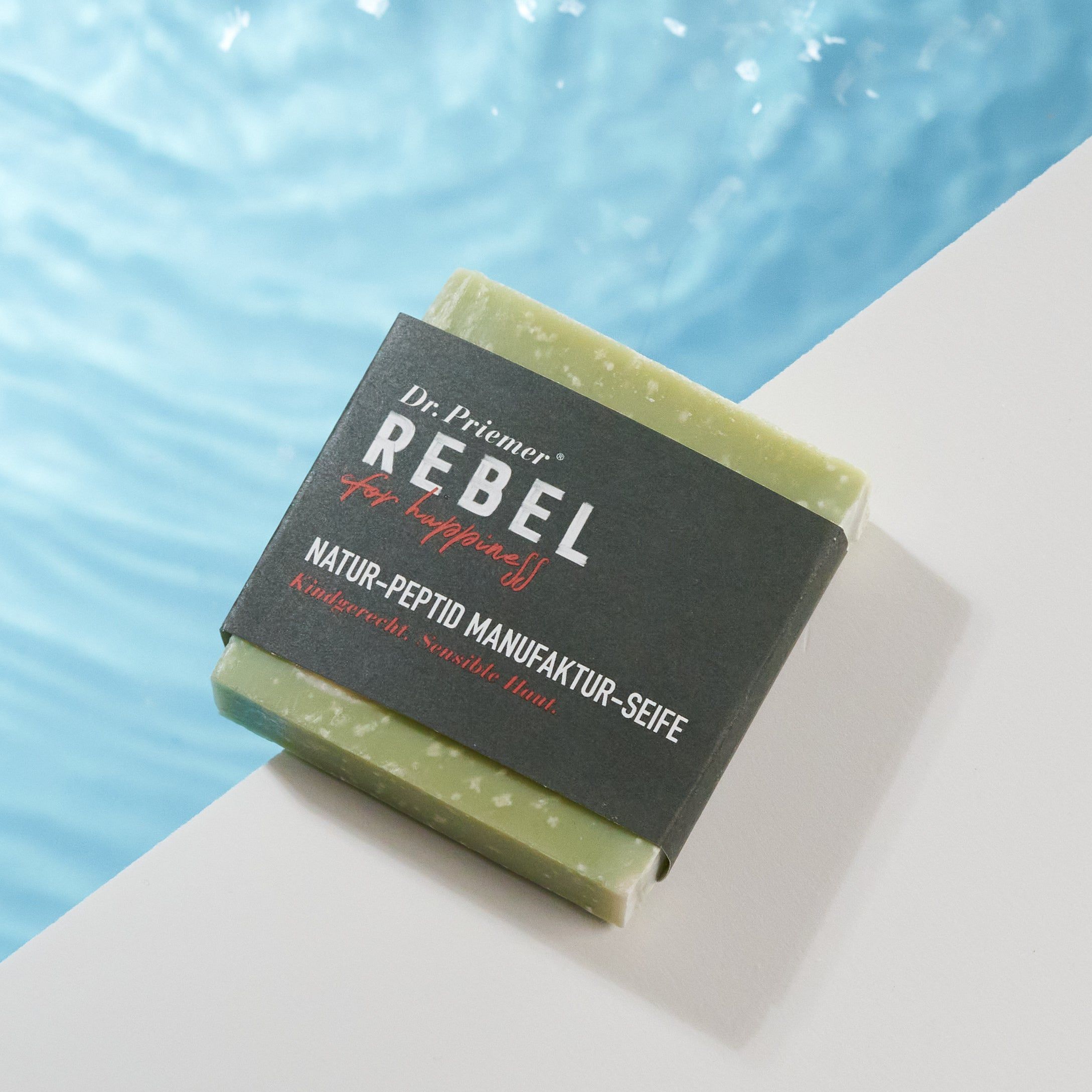Seife vor blauem Wasserhintergrund. Aufschrift: Dr. Priemer, REBEL for happiness, Natur-Peptid Manufaktur-Seife.