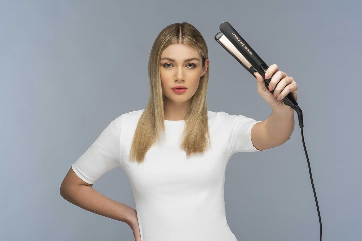 Blonde Frau hält ein schwarzes Glätteisen hoch. Sie blickt in die Kamera. Das Glätteisen ist zu sehen.