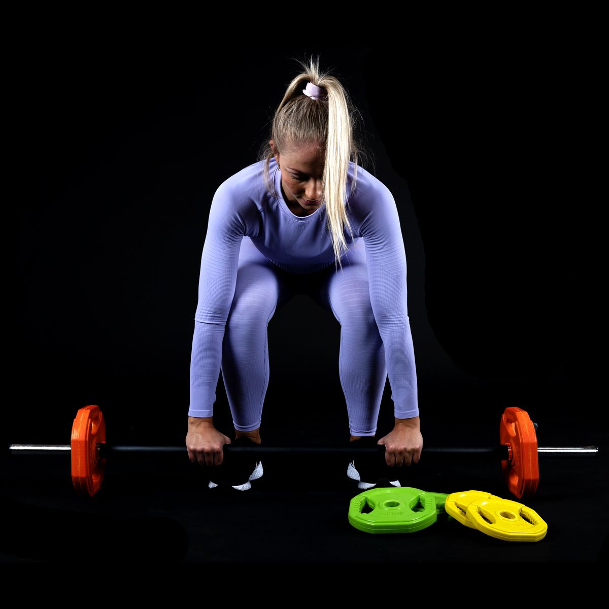 Frau in Sportkleidung mit Langhantel. Orange, grüne und gelbe Gewichtsscheiben. Zwei Scheiben liegen.