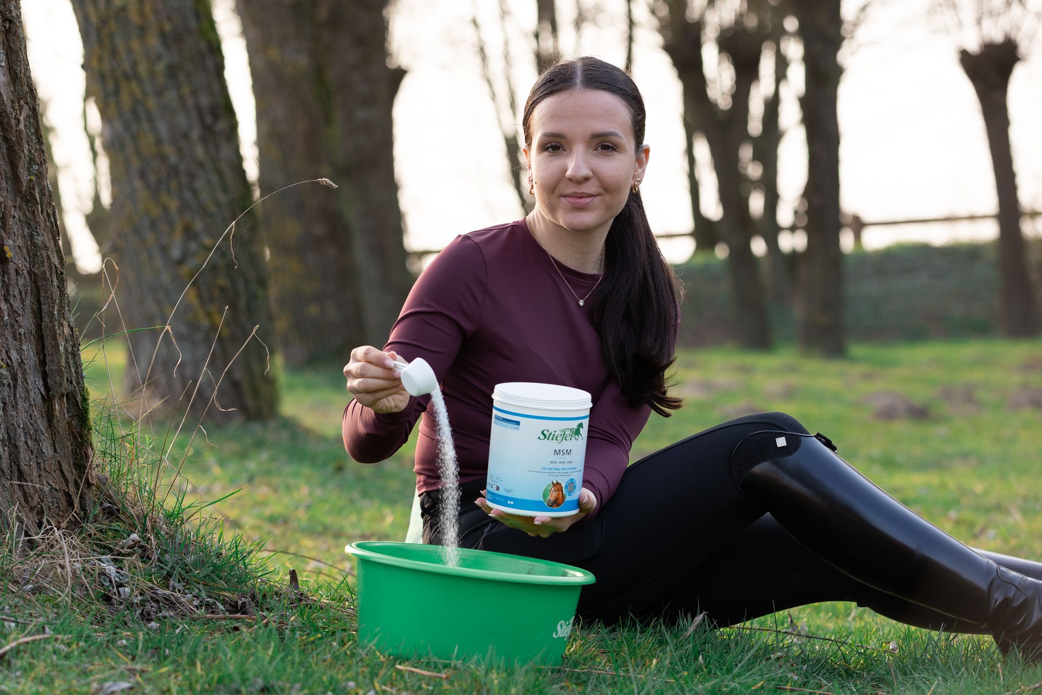 Frau sitzt im Freien und hält einen Behälter und einen Löffel. Sie schüttet Pulver in einen grünen Behälter. Produkt: Stiefel MSM.