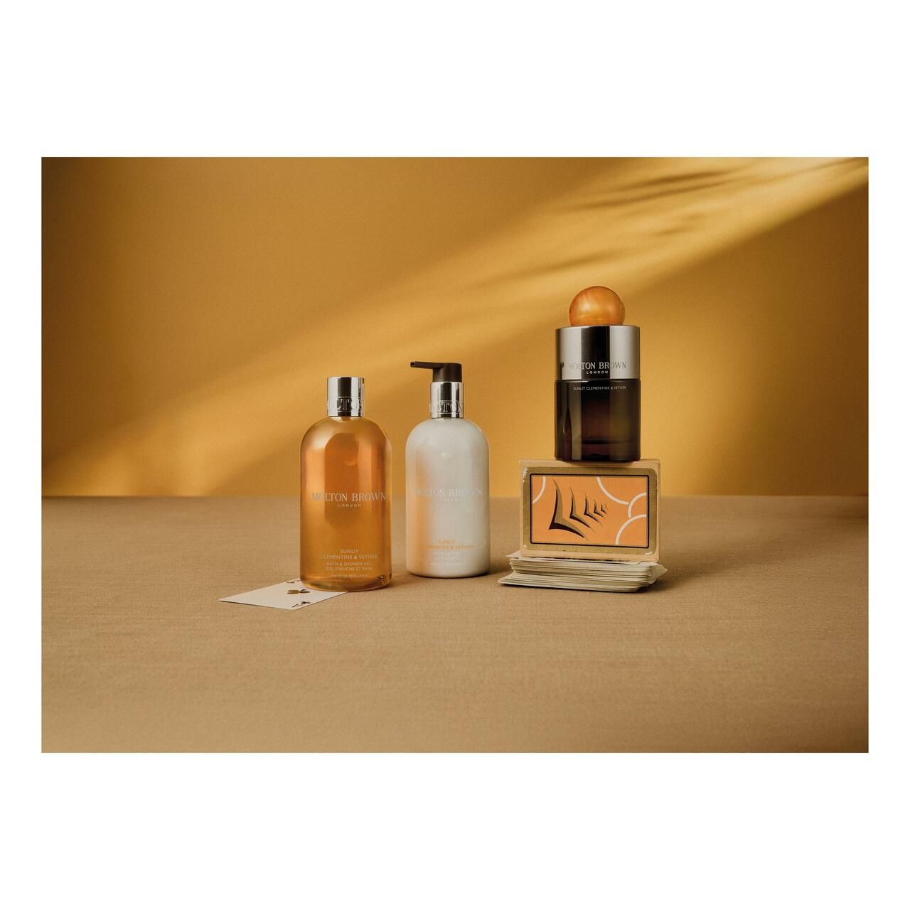 Drei Produkte auf einer beigen Oberfläche. Links: Duschgel. Mitte: Lotion. Rechts: Parfum. Aufschrift: Molton Brown.