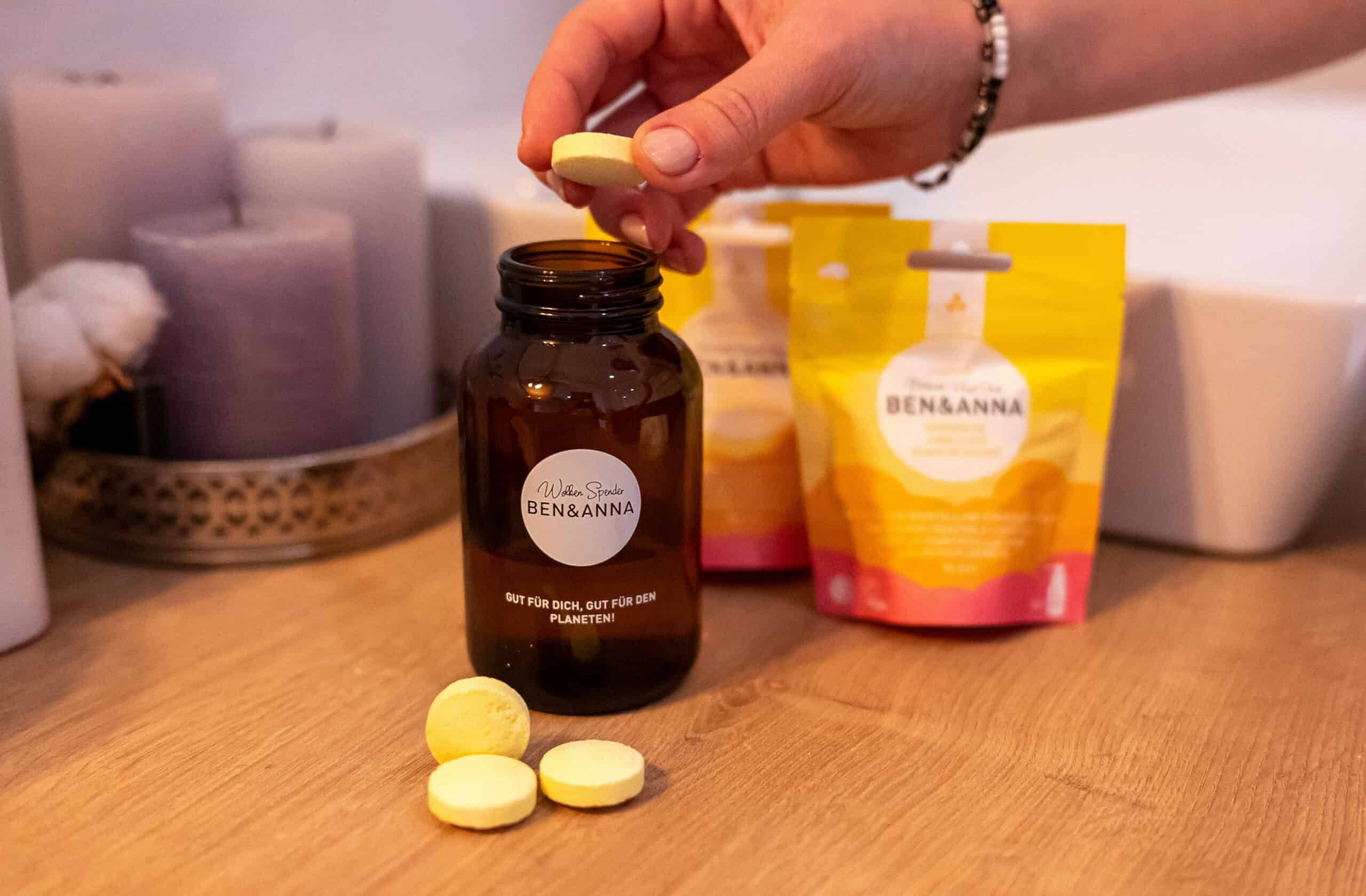 Braune Glasflasche und gelbe Tabletten. Aufschrift: Ben & Anna. Gelbe Tabletten werden aus der Flasche entnommen. Im Hintergrund: Verpackung.
