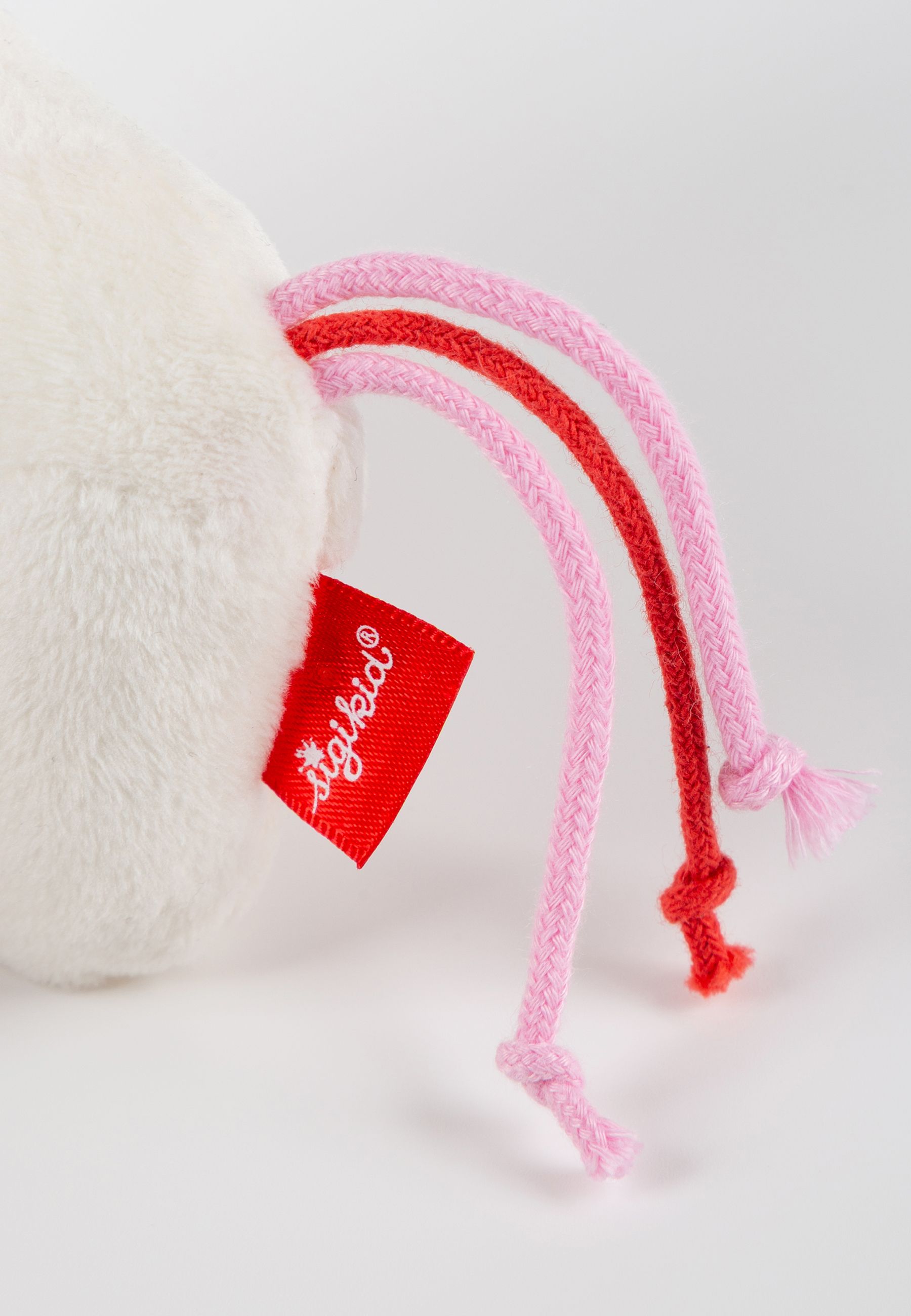 Rückansicht eines weißen Stoffeinhorns. Rotes Etikett mit SIGIKID-Logo. Rosa und rote Kordeln.