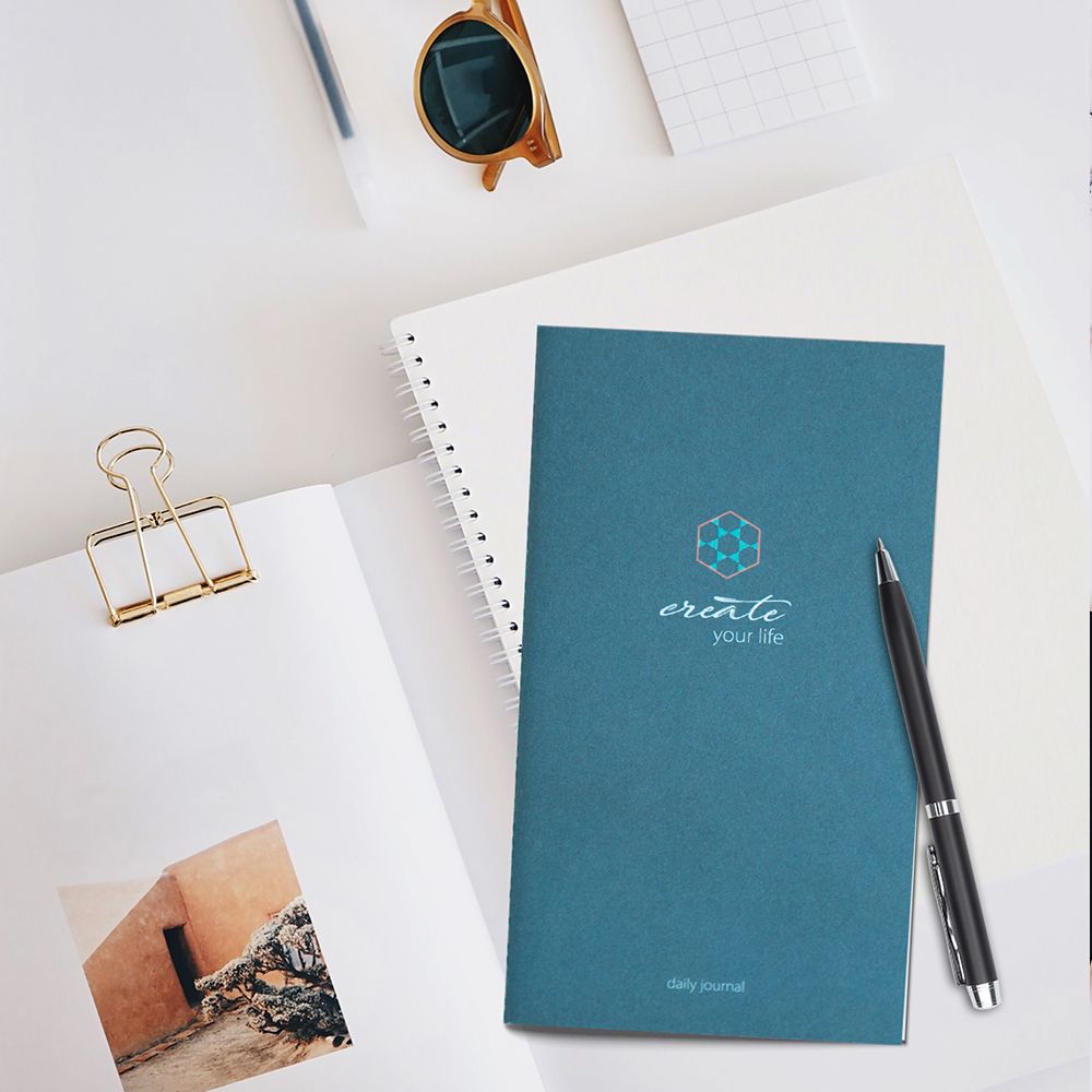 Blaues Tagebuch mit Stift. Aufschrift: create your life, daily journal. Daneben Brille und Notizblock.