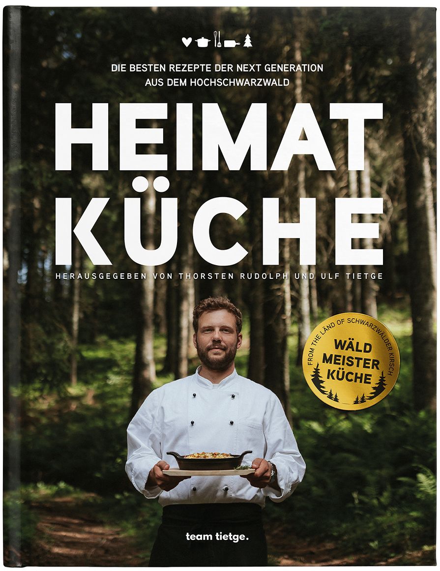 Kochbuch "HEIMAT KÜCHE". Titel in Großbuchstaben. Auf dem Cover ein Mann mit Kochjacke, der ein Gericht präsentiert. Im Hintergrund Wald.