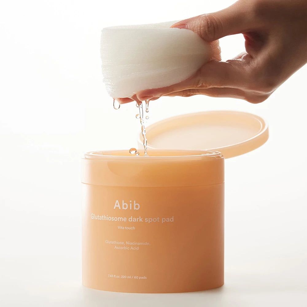 Beige Dose mit Deckel, Pads und Flüssigkeit. Hand hält Pads über der Dose. Aufschrift: Abib Glutathiosome dark spot pad.