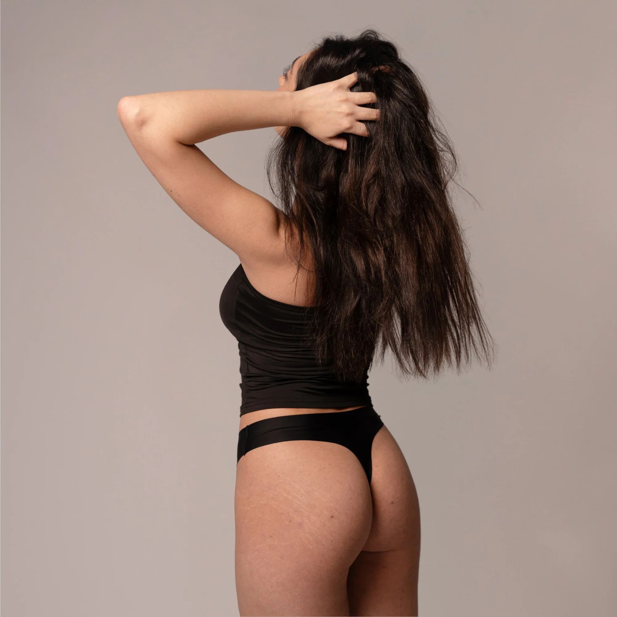 Frau in schwarzem Top und Slip, von hinten. Sie berührt ihre Haare.