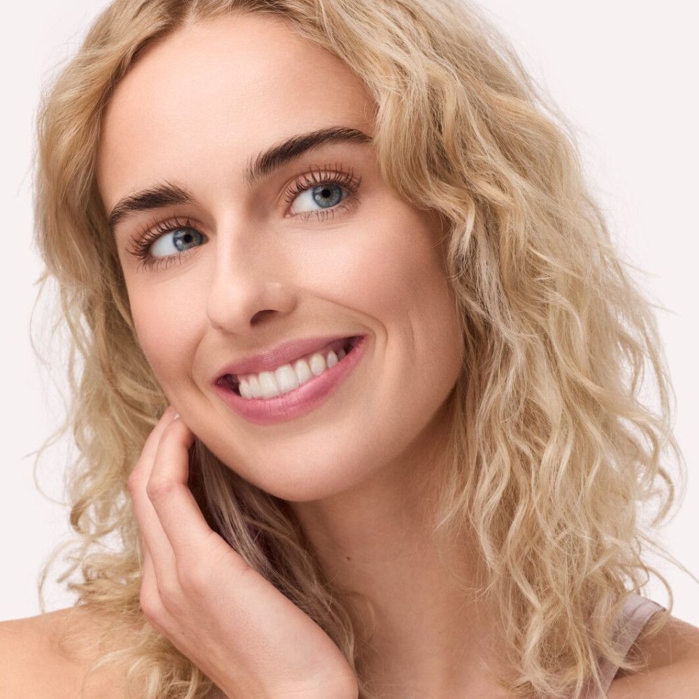 Porträt einer Frau mit blonden Locken. Sie lächelt und berührt ihre Wange. Blaue Augen.