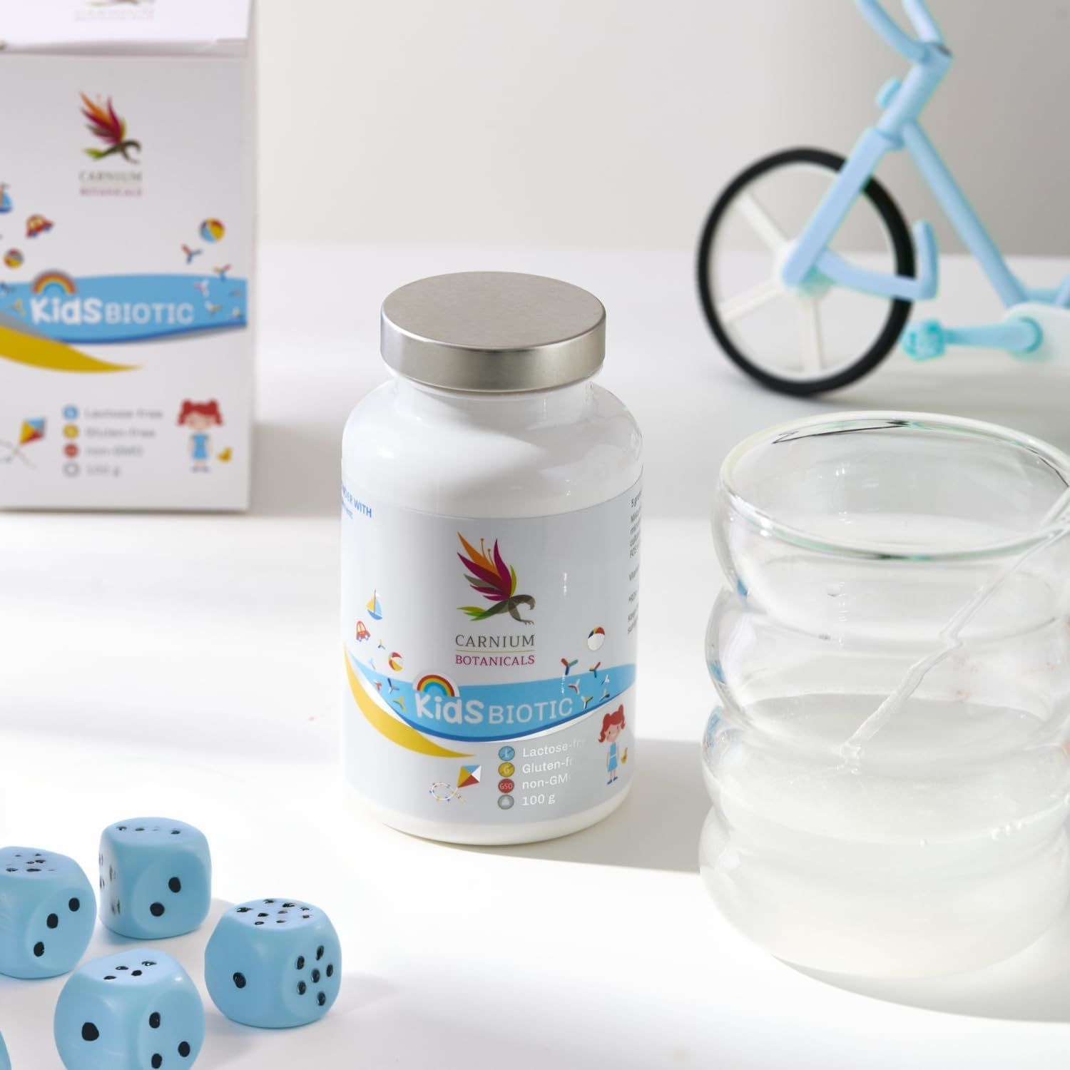 Weiße Flasche mit silbernem Deckel. Aufschrift: Kids Biotic, Carnium Botanicals. Würfel, Glas und Spielzeugfahrrad.