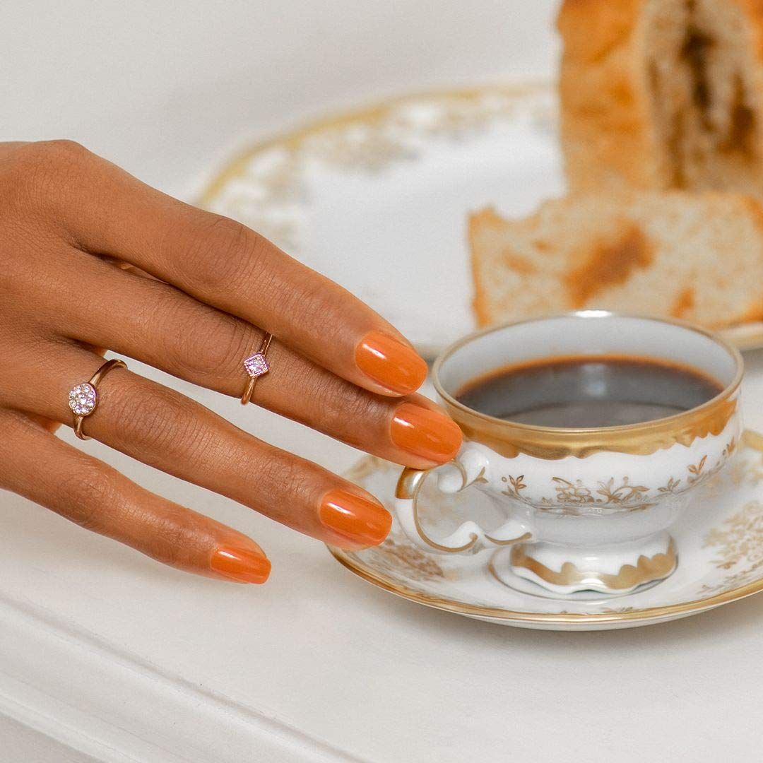 Hand mit orange lackierten Nägeln neben einer Kaffeetasse und Gebäck. Ringe an den Fingern.