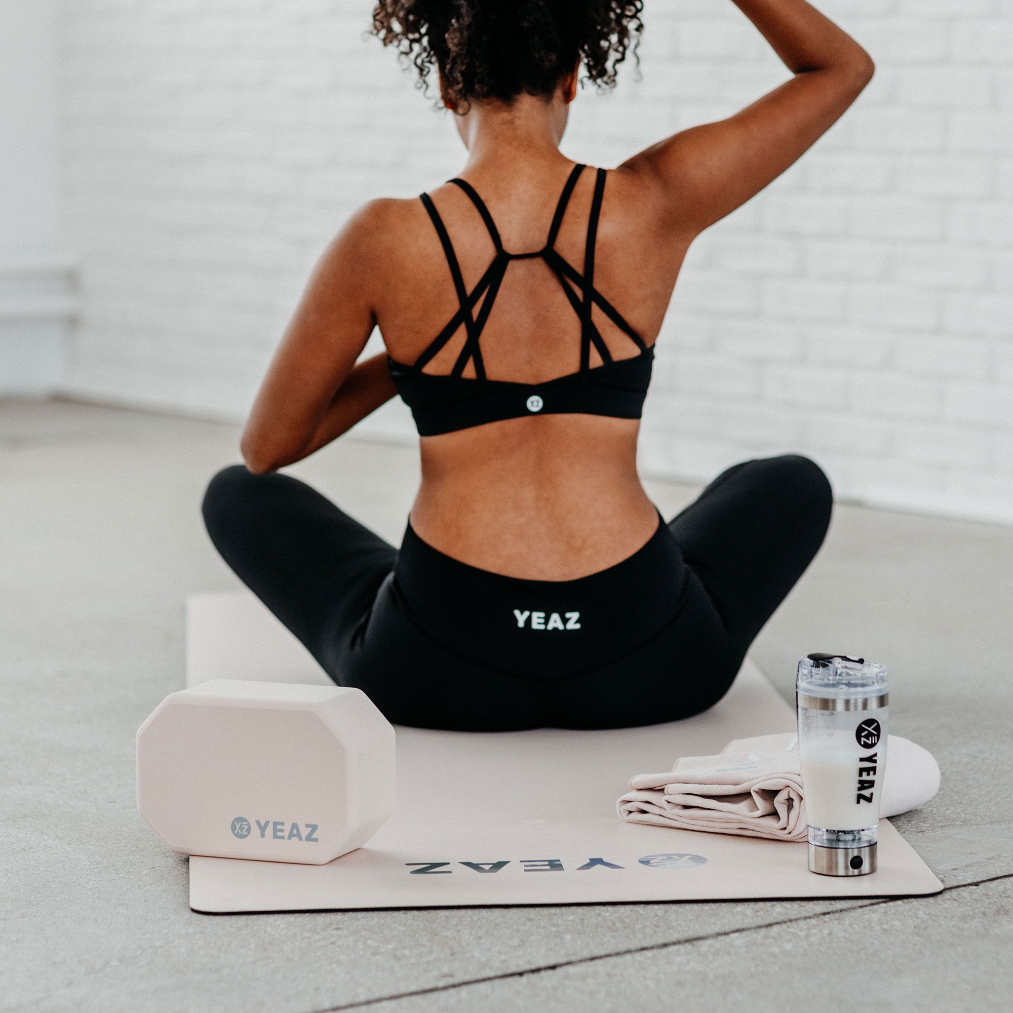 Eine Frau in Sportkleidung übt Yoga neben einem hellrosa YEAZ Yoga-Block, einer Matte und einer Wasserflasche.