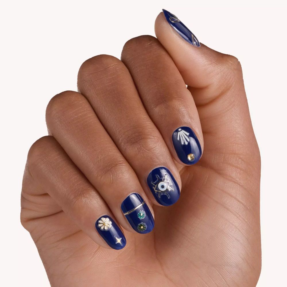 Hand mit lackierten Nägeln. Blaue Nägel mit goldenen und weißen Nagelstickern. Motive: Auge, Blüte, Muschel.