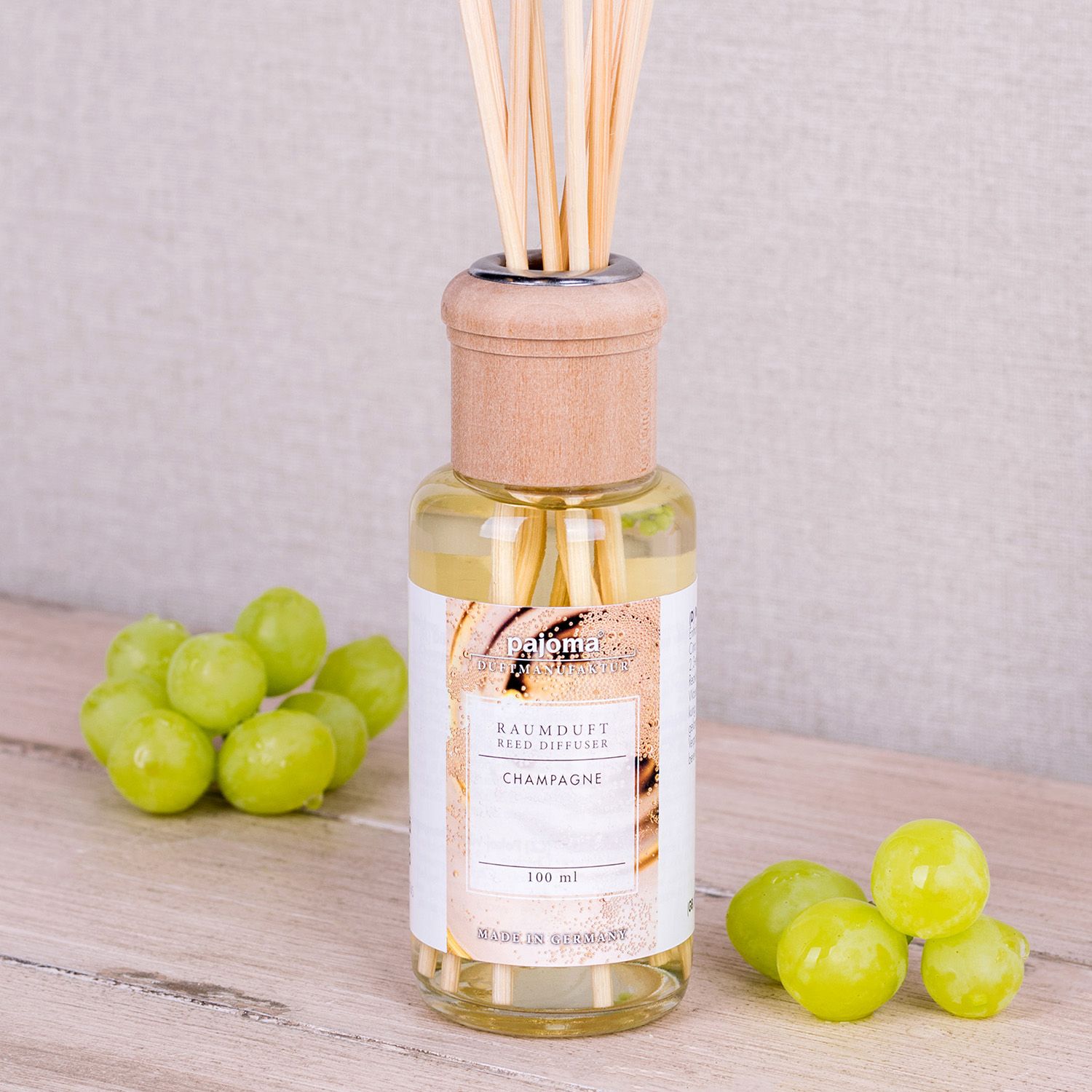 Glasflakon mit Holzverschluss und Duftstäbchen. Aufschrift: Champagne. Neben dem Flakon liegen grüne Trauben.
