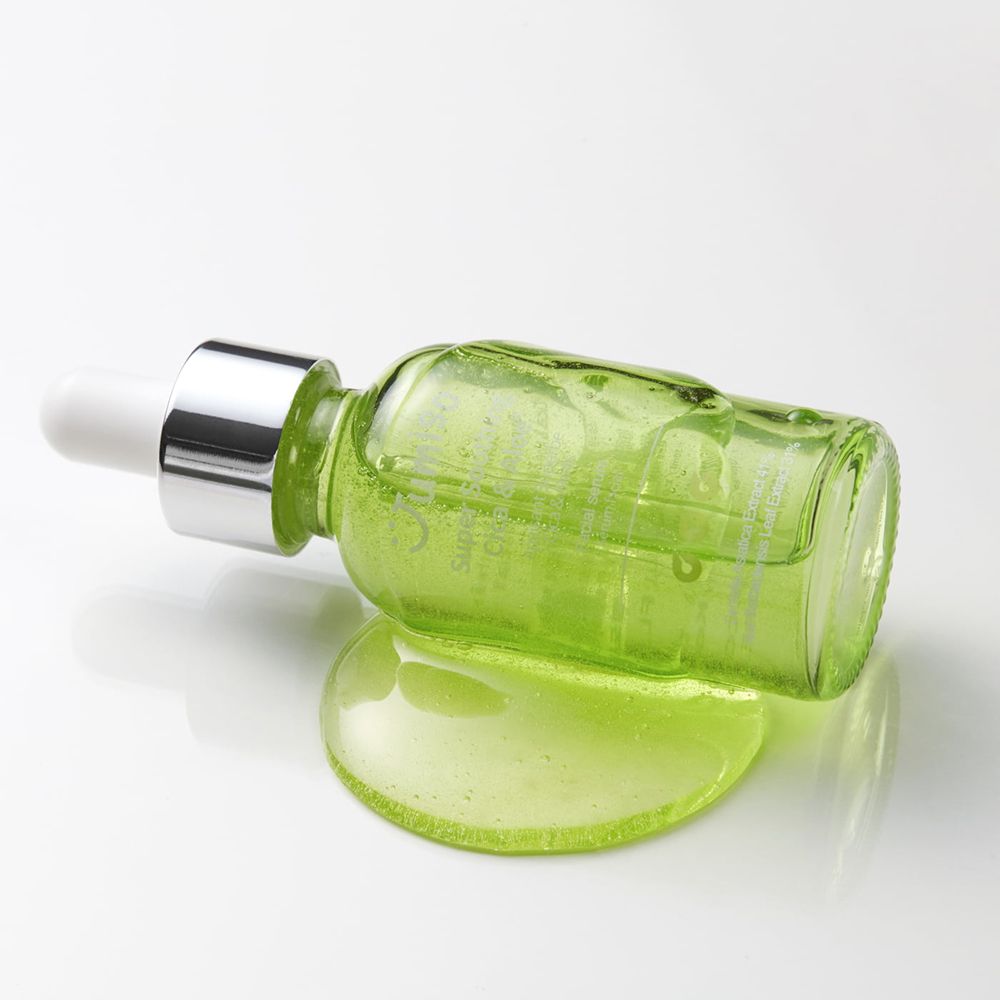 Grüne Glasflasche mit weißem Deckel und Pipette liegt auf einer weißen Oberfläche. Daneben eine Serum-Pfütze.