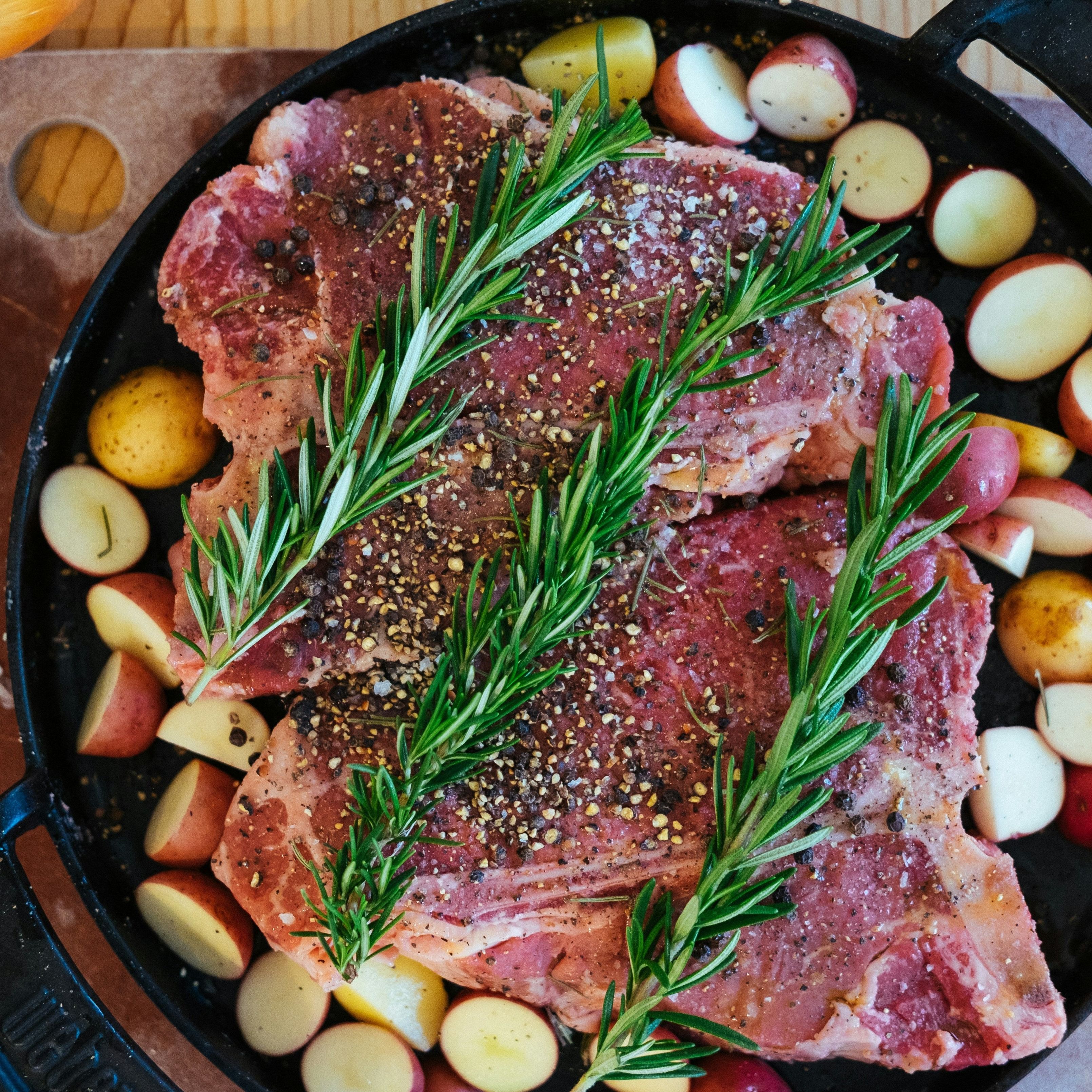 Zwei Steaks mit Rosmarinzweigen und Pfeffer auf Bratpfanne. Kleine Kartoffeln umgeben das Fleisch.