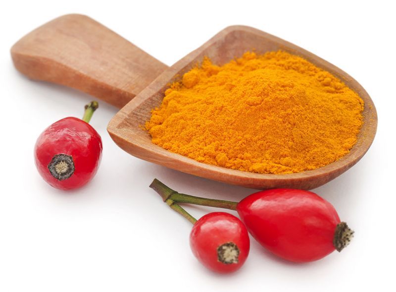 Holzlöffel mit orangefarbenem Pulver, daneben drei Hagebutten. Weißer Hintergrund. Produkt nicht identifizierbar.
