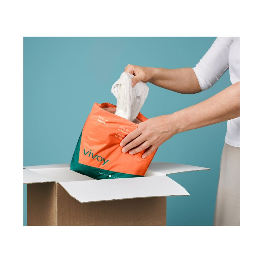 Hände entnehmen Vivoy Inkontinenz Pants aus einer Verpackung. Orange-grüne Verpackung. Hintergrund: hellblau und eine geöffnete Box.