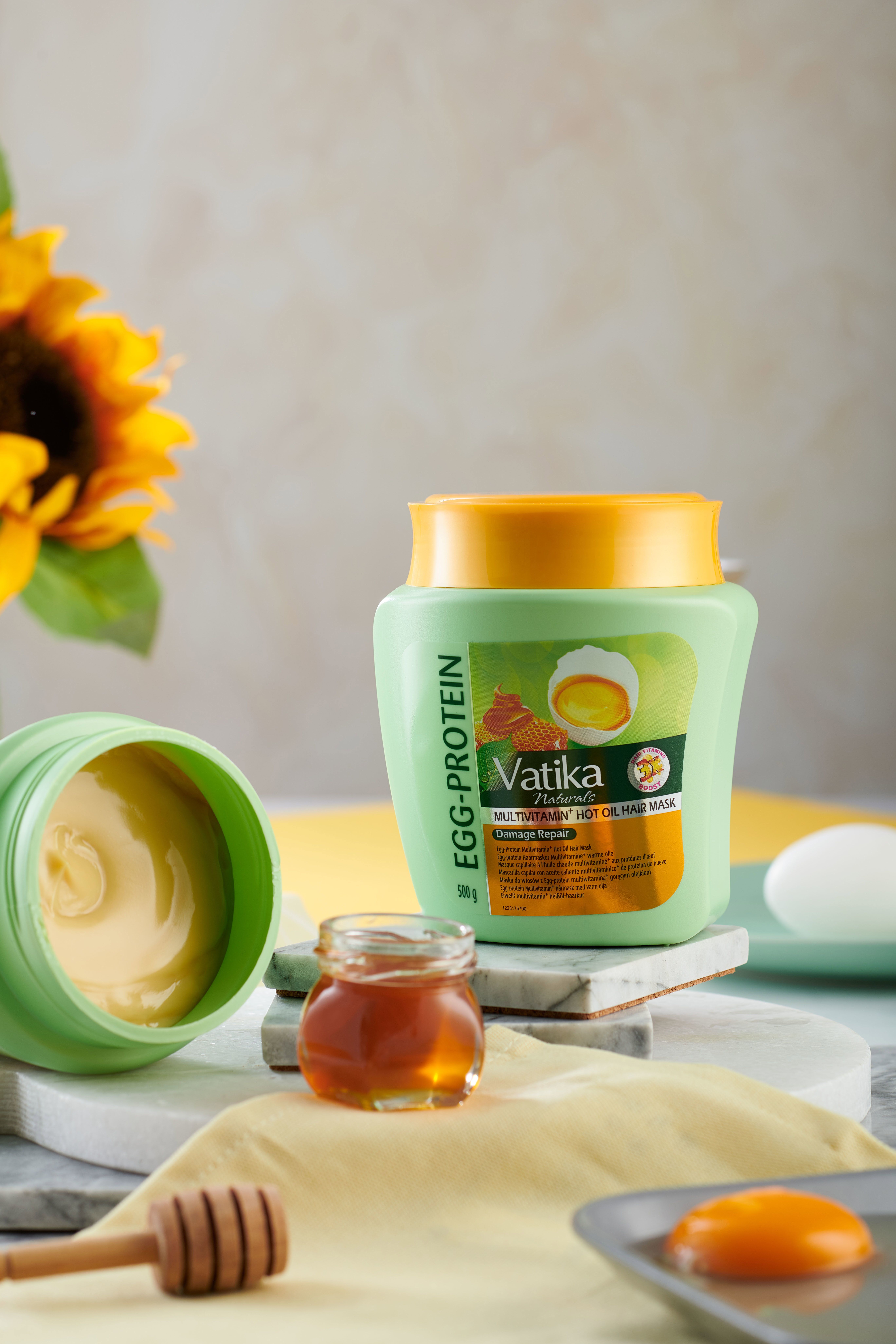 Geöffnete grüne Haarmaske mit gelbem Deckel. Honigglas, Eierschalen, Holzstab. Text: Vatika Naturals, Egg-Protein, Multivitamin, Hot Oil Hair Mask.