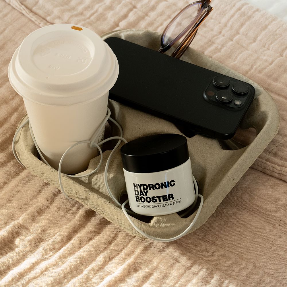 Becher, Smartphone und Tiegel mit Aufschrift: HYDRONIC DAY BOOSTER, Vegan CBD Day Cream + SPF 20, in einem Tablett.