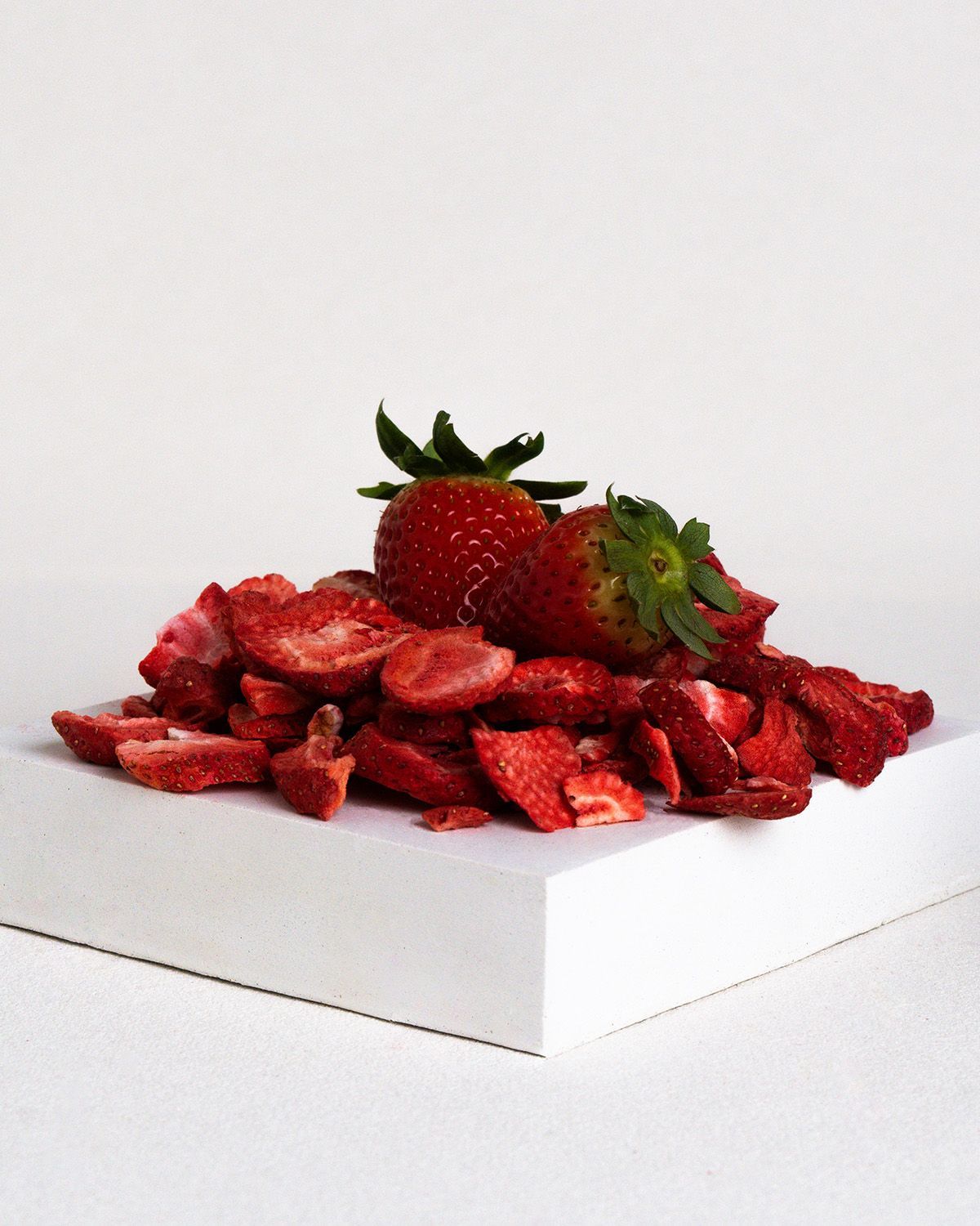 Haufen gefriergetrockneter Erdbeerscheiben und zwei frische Erdbeeren. Auf weißem Untergrund.