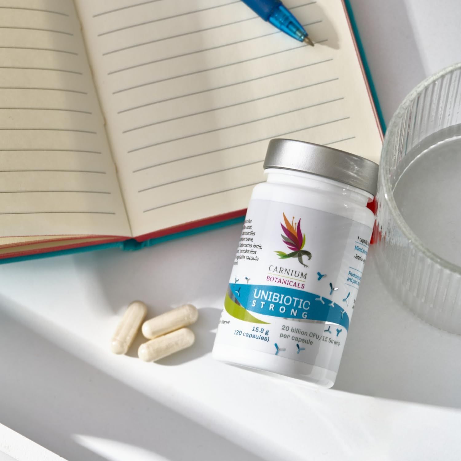 Flasche mit Kapseln neben Notizbuch und Glas. Aufschrift: Unibiotic STRONG, 20 Milliarden CFU/15 Stämme. Marke: CARNIUM BOTANICALS.