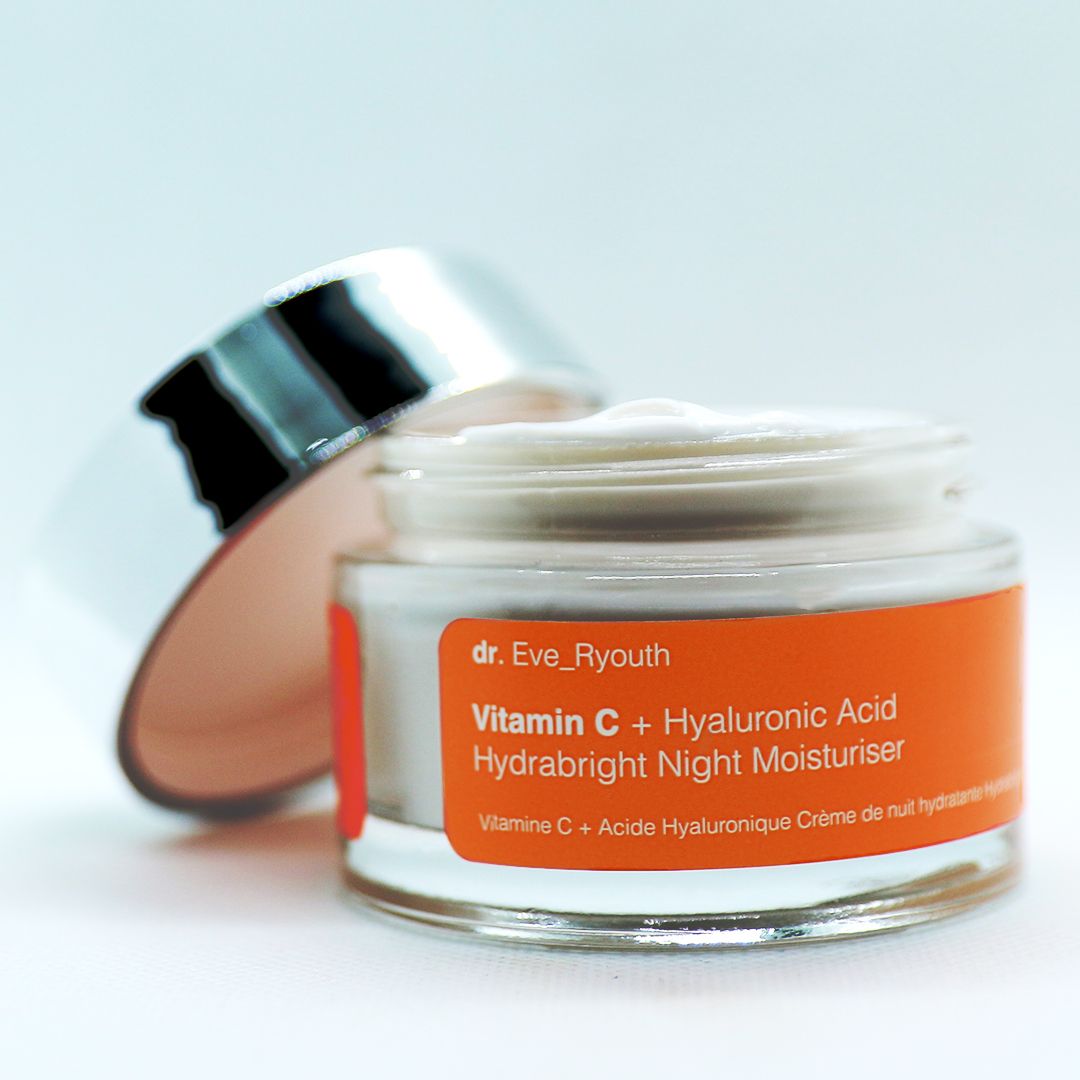 Geöffneter Creme-Tiegel mit silbernem Deckel. Das Innere zeigt eine weiße Creme. Orangefarbenes Etikett mit Produktinformationen.