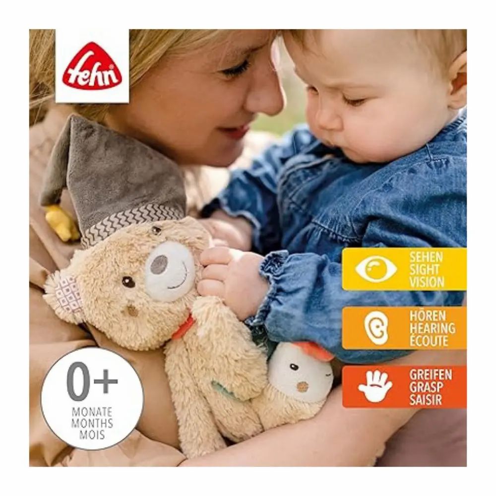 Baby hält Stofftier mit Mütze, rotem Schal, Stern und Fisch-Anhänger. Marke: fehn. Mit Text und Symbolen.