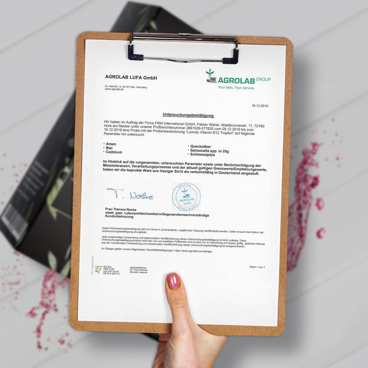 Person hält Klemmbrett mit Dokument. Aufschrift: Agrolab LUFA GmbH, Unterschungsbericht. Logo: Agrolab Group. Teilweise Produktverpackung im Hintergrund.
