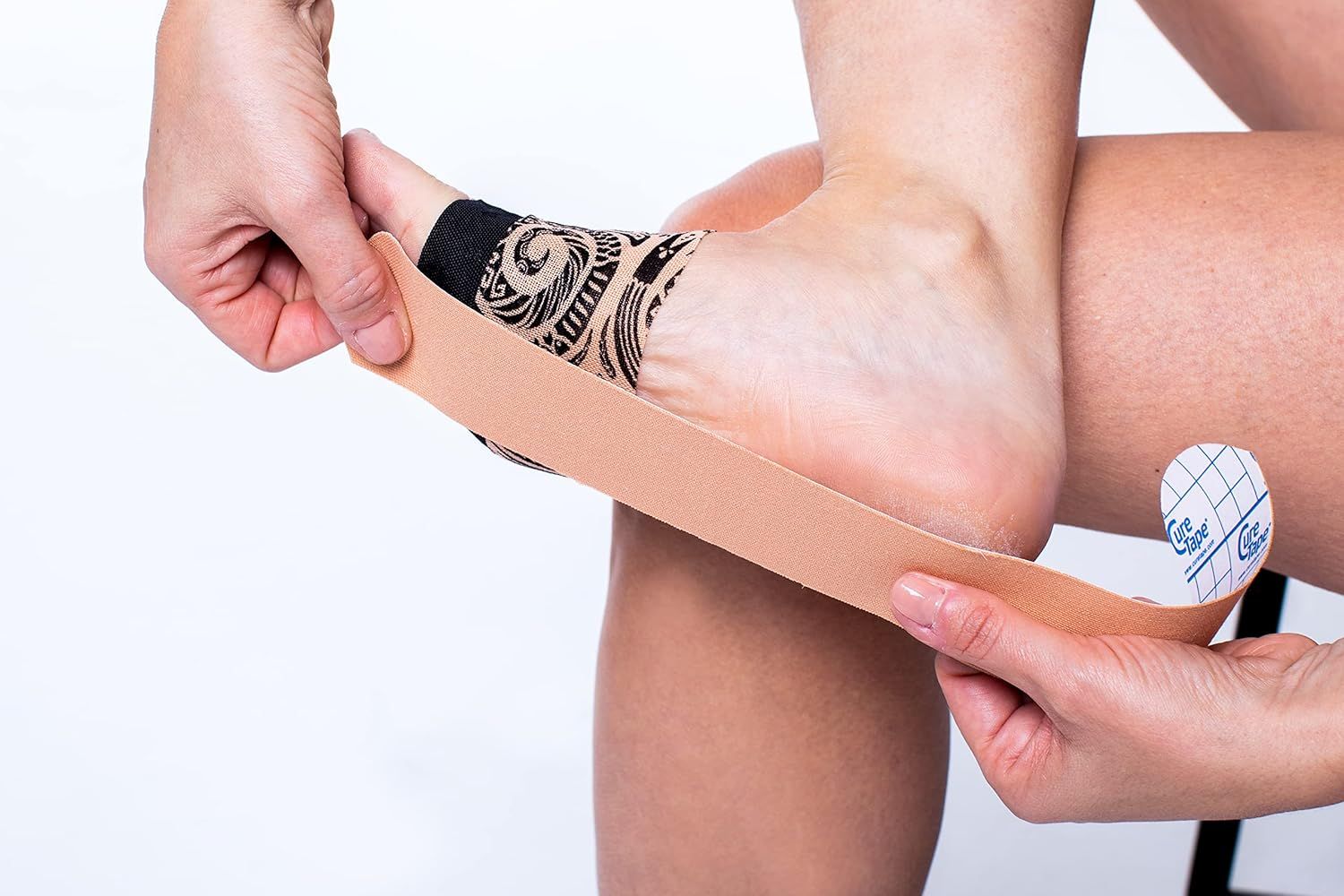 Person klebt beige Kinesiologie-Tape auf Fuß. Tape wird auf Fuß geklebt. Person sitzt. Tattoo am Fuß.