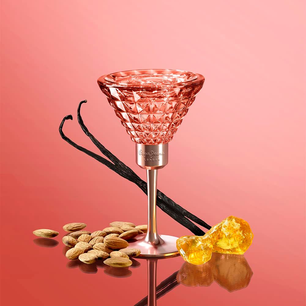 Parfümflakon in Kelchform. Rosa Glas mit geometrischem Muster. Vanilleschoten, Mandeln und bernsteinfarbene Stücke liegen daneben.