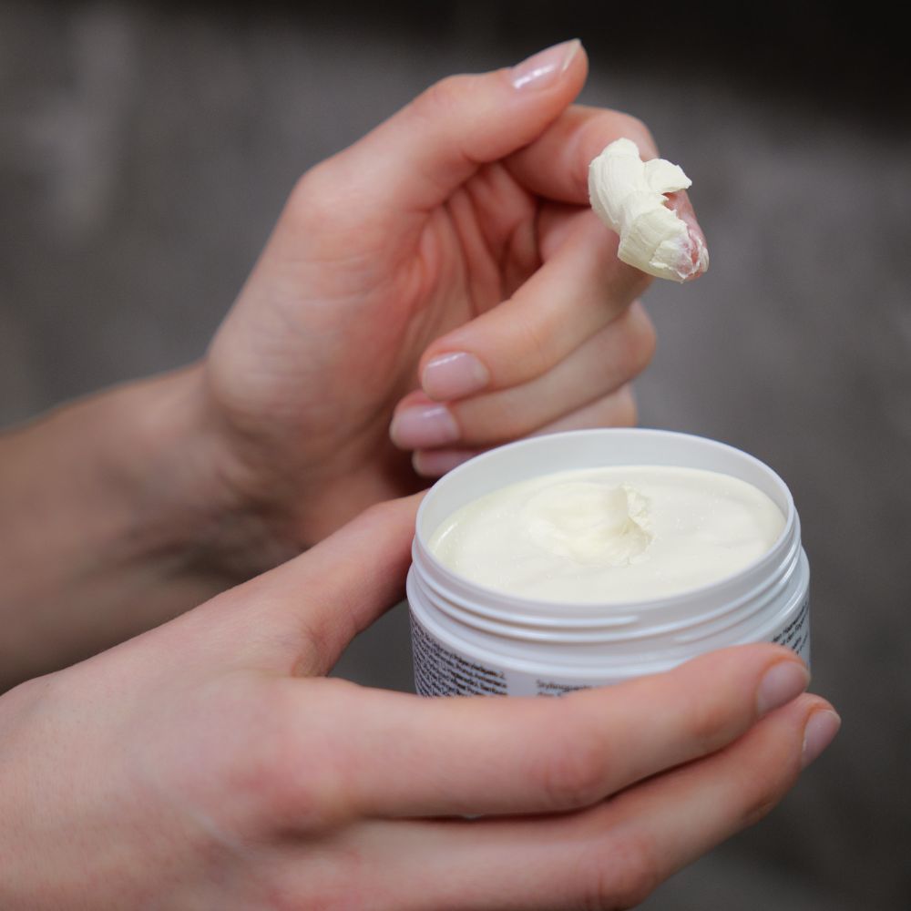 Hände mit Genki Styling Paste. Grauer Deckel, schwarzer Schriftzug, gelber Farbfleck. Creme.