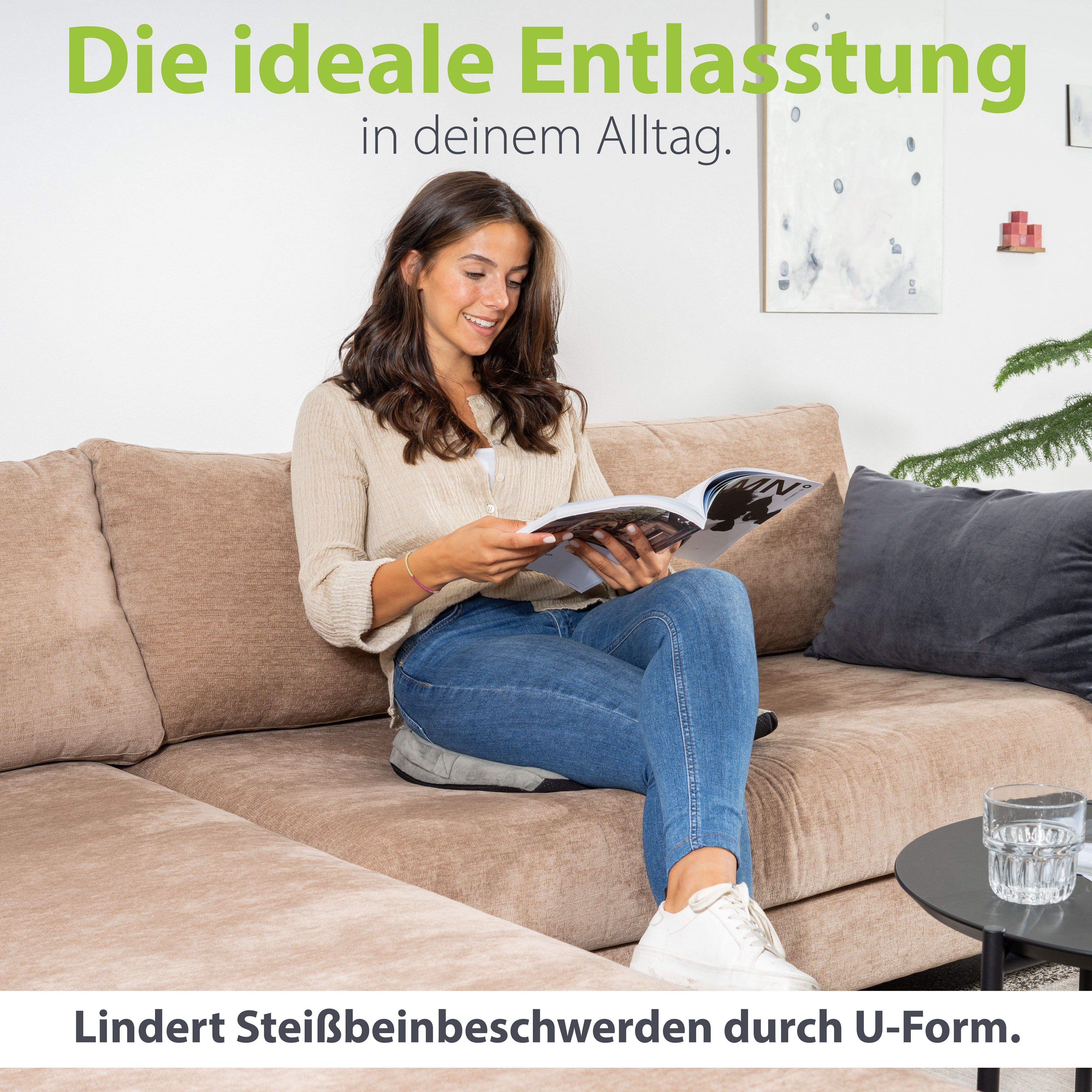 Frau sitzt auf Sofa mit Keilkissen. Text: Lindert Steißbeinbeschwerden durch U-Form. Kissen ist sichtbar.