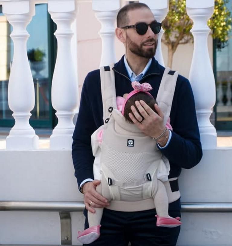Ein Mann trägt ein Baby in einer beige Babytrage. Das Baby schaut nach innen.
