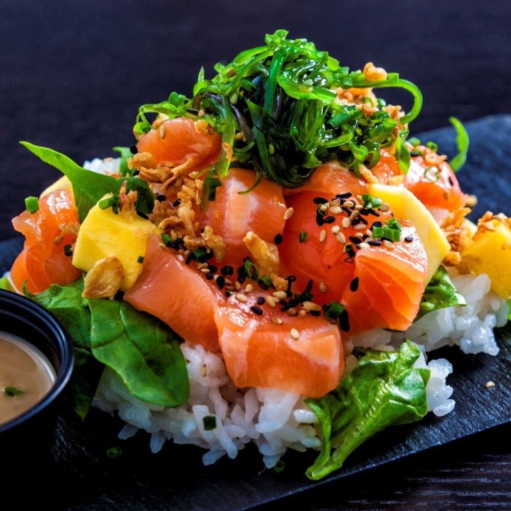 Gericht mit Lachs, Reis, Algen, Mango und Sesam. Schwarzer Teller, kleine Schale mit Sauce.