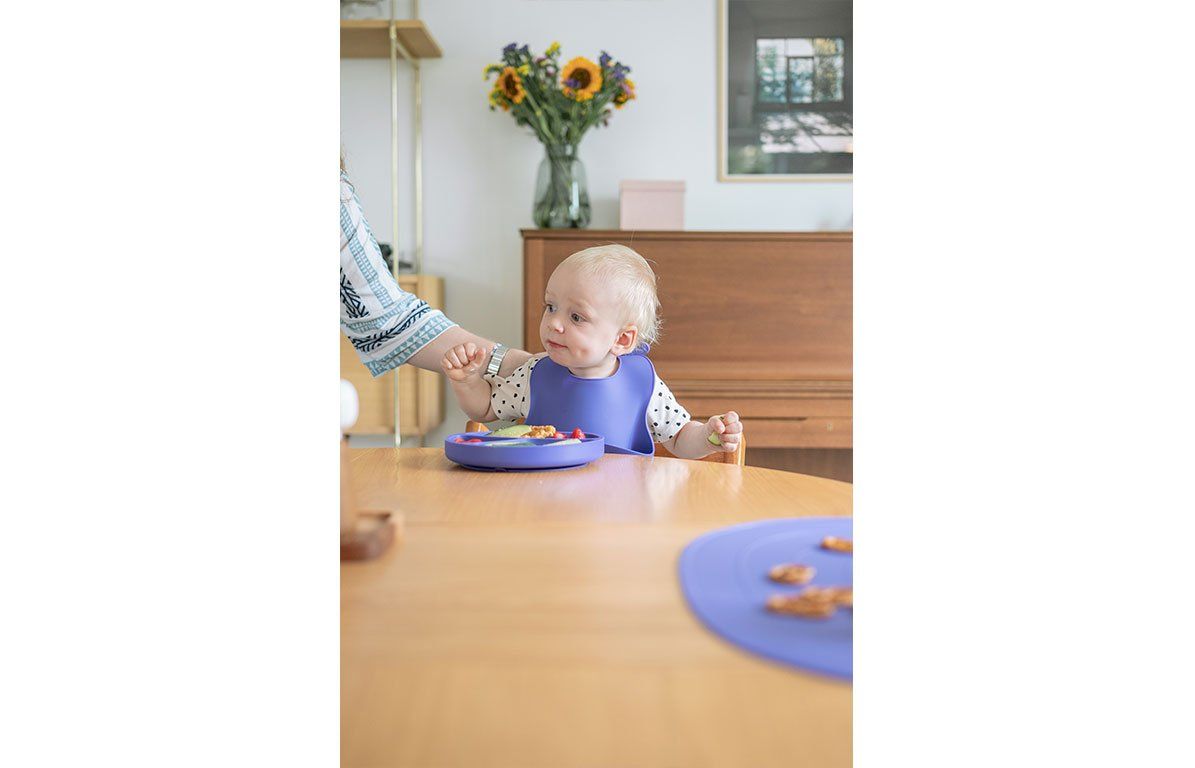Baby am Tisch mit lila Lätzchen. Person reicht Essen. Auf dem Tisch Teller mit Essen.