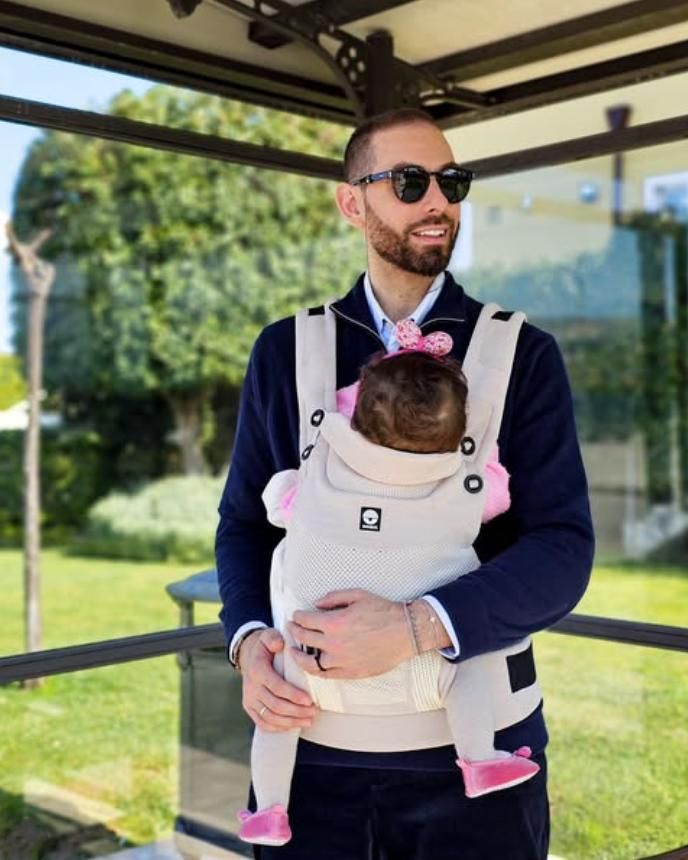 Ein Mann trägt ein Baby in einer beige Babytrage. Das Baby schaut nach innen.