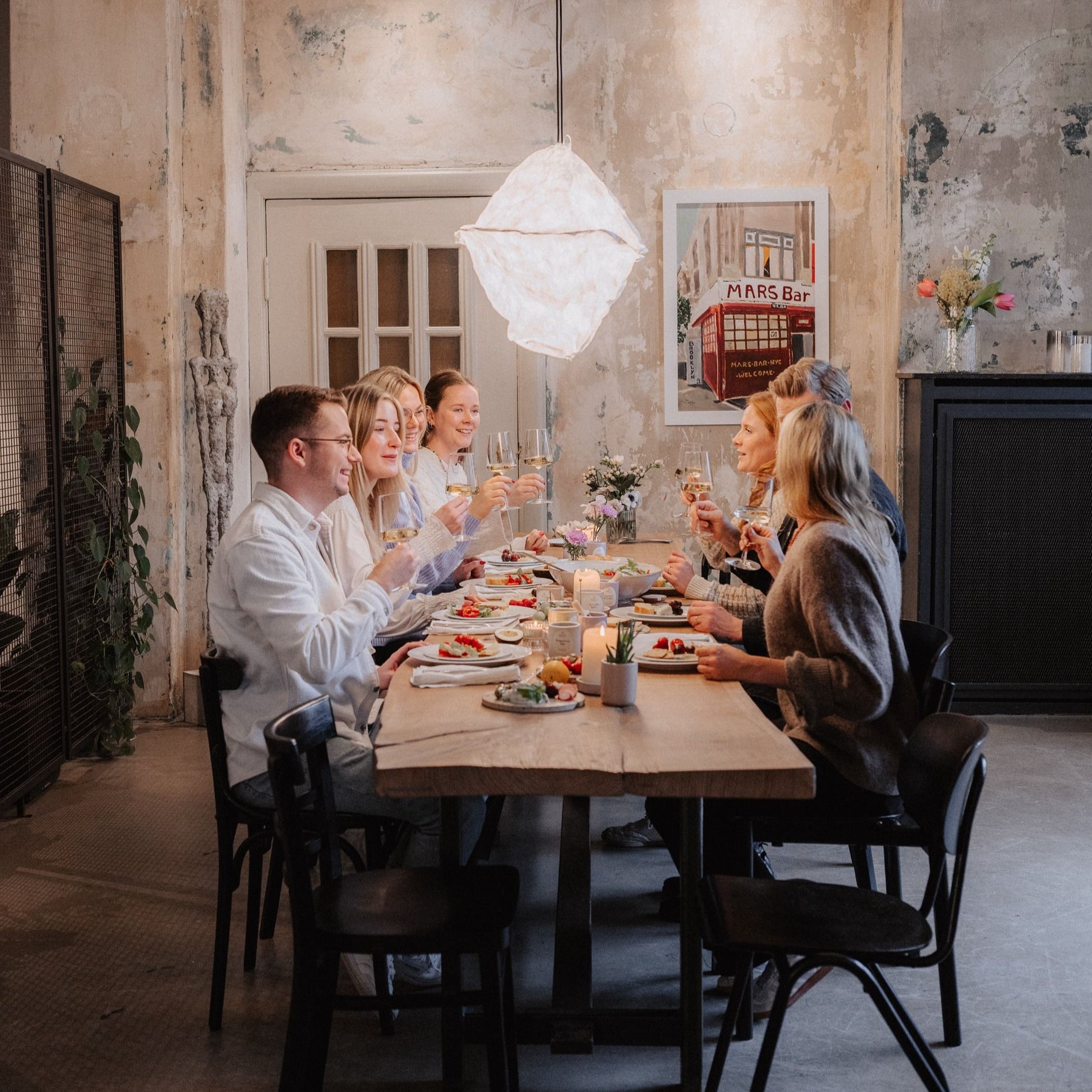 Gruppe von Personen sitzt an einem Holztisch. Sie stoßen mit Weingläsern an. Essen und Kerzen auf dem Tisch.