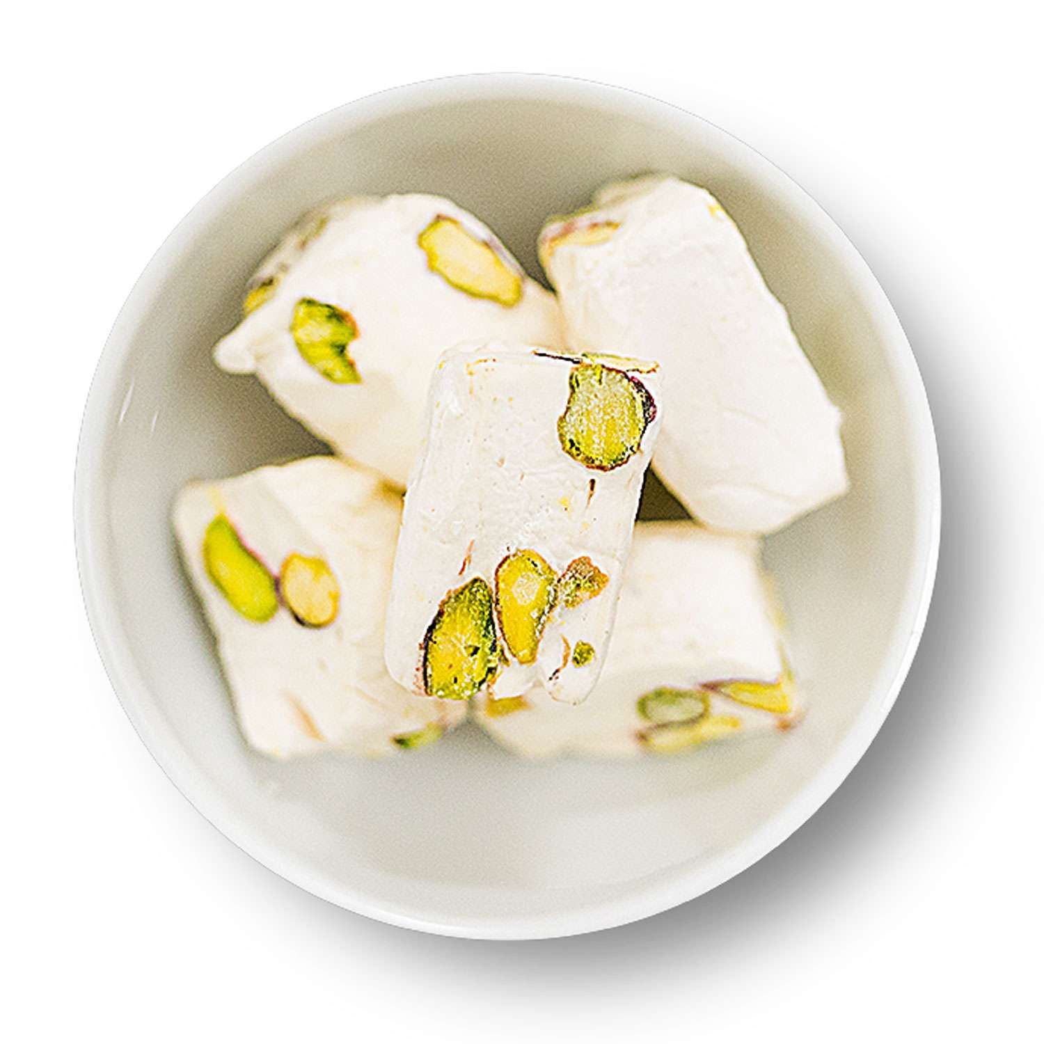 Weißes Nougat mit Pistazien in einer weißen Schale. Mehrere Stücke, rechteckig und unregelmäßig geformt.