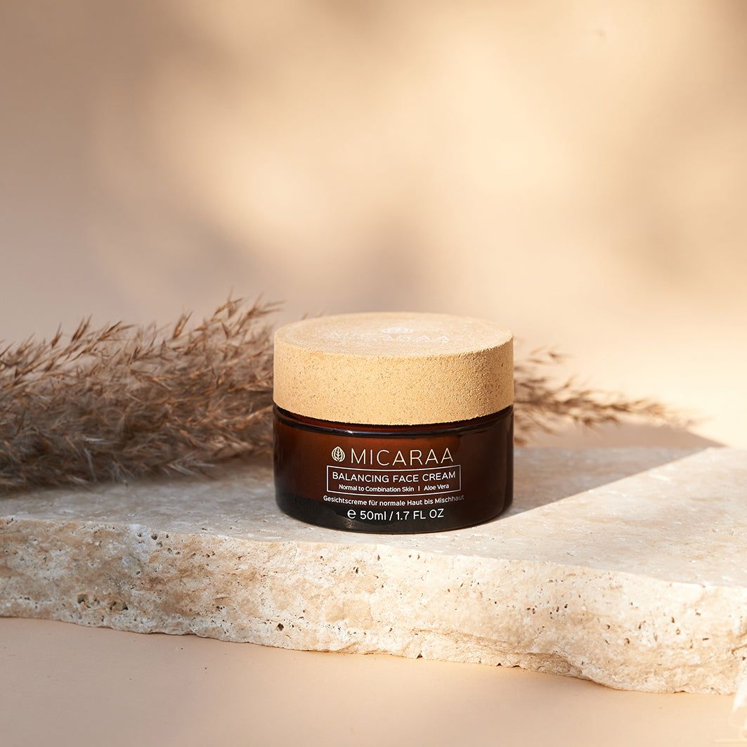 Braune Creme-Dose mit Holzdeckel auf Stein. Aufschrift: MICARAA Balancing Face Cream, Aloe Vera. 50ml/1.7 FL OZ. Hintergrund: Pflanze.