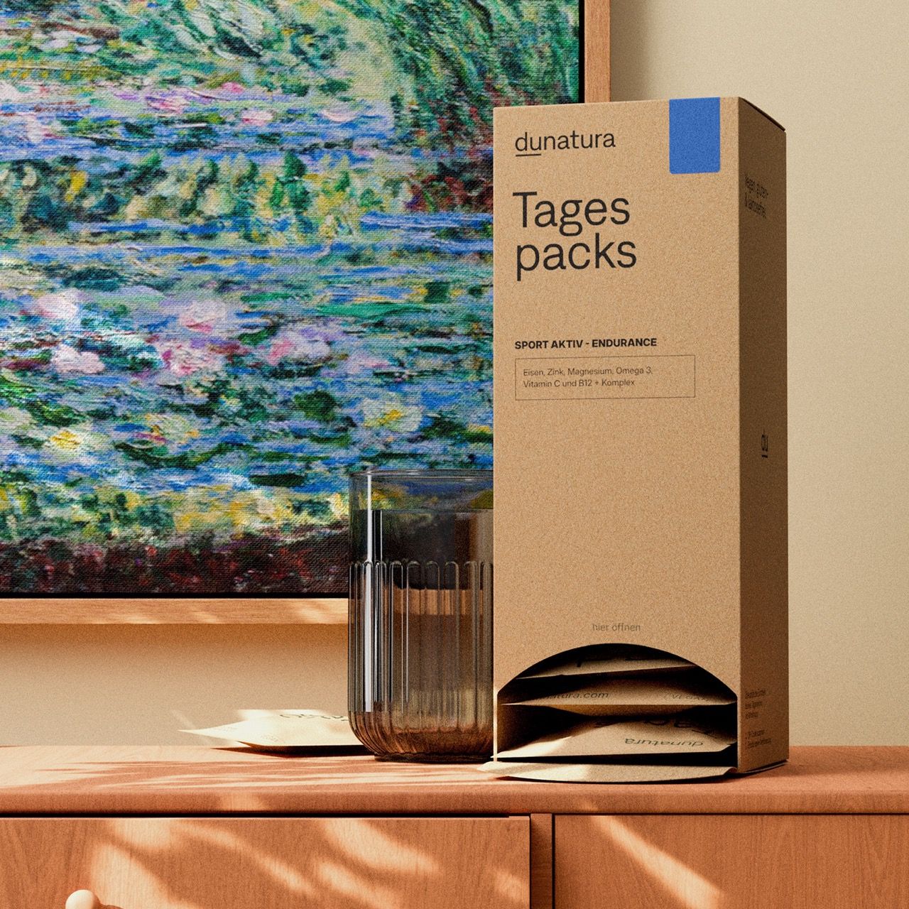 Produktverpackung 'dunatura Tagespacks Sport Aktiv Endurance' auf einem Tisch. Glas Wasser daneben. Kunstwerk im Hintergrund.