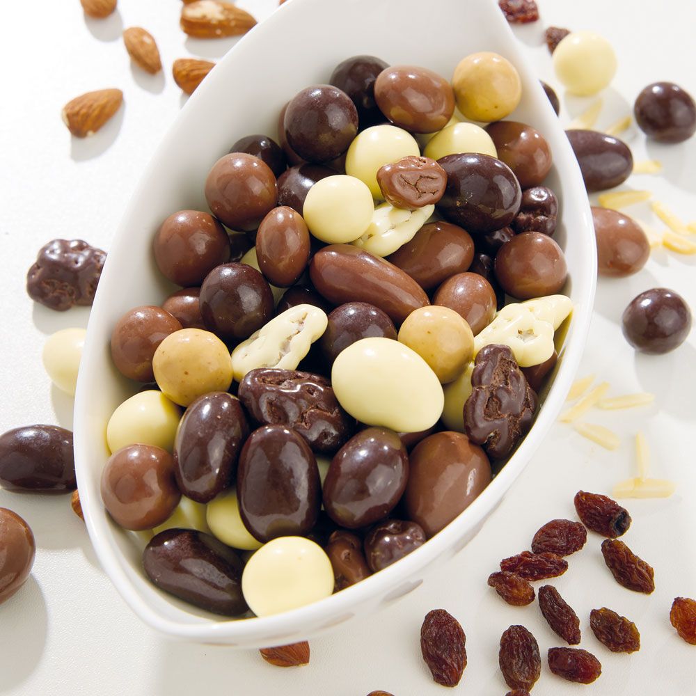 Weiße Schale gefüllt mit Schokoladen-Knabbermischung. Verschiedene Formen und Farben. Mandeln und Rosinen daneben.